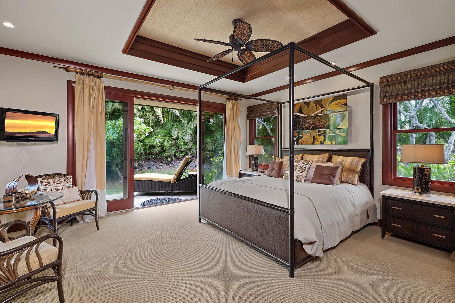 This is a primary bedroom featuring a four-poster bed with neutral bedding, complemented by decorative pillows. The room has a tropical aesthetic with a ceiling fan, woven ceiling detail, and access to an outdoor patio through sliding glass doors. A flat-screen TV is mounted on the wall, and the room is furnished with rattan seating and a dark wood nightstand with a lamp.