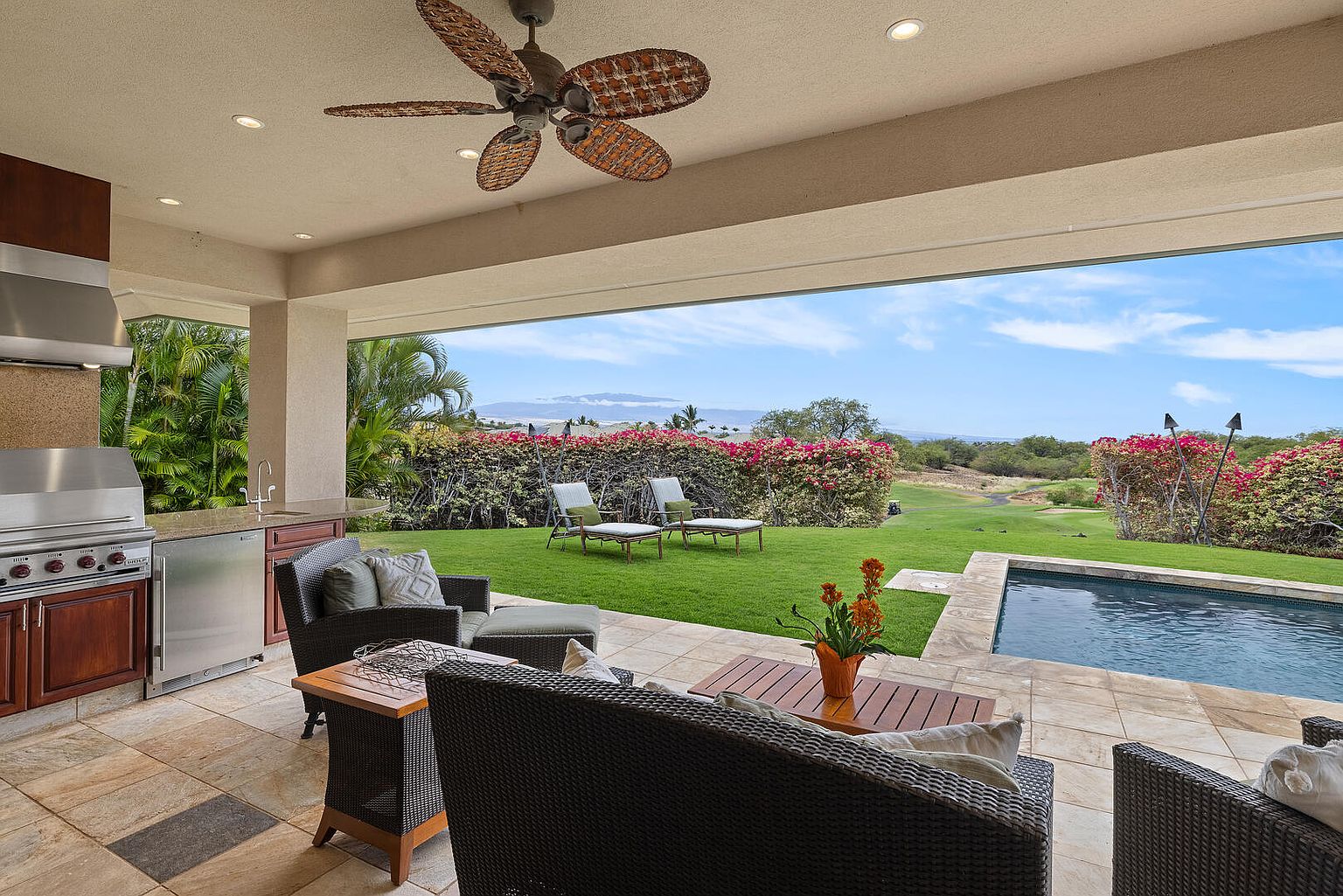 This image showcases a luxurious outdoor patio area, seamlessly blending indoor and outdoor living. The patio features comfortable wicker furniture, a built-in outdoor kitchen with stainless steel appliances, and a view of a well-manicured lawn, a swimming pool, and distant mountains. The space is covered, providing shade and protection from the elements, while the open design allows for natural light and airflow.