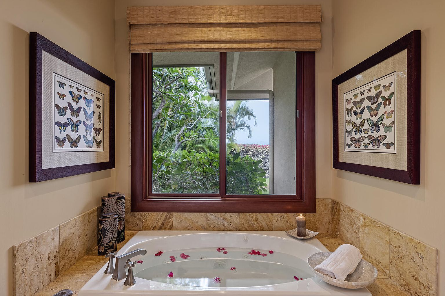 This is a luxurious primary bathroom featuring a soaking tub filled with water and rose petals, positioned beneath a window offering a view of lush greenery. The walls are a neutral tone, complemented by marble countertops and framed butterfly artwork on either side of the window. The overall impression is one of relaxation and tranquility.