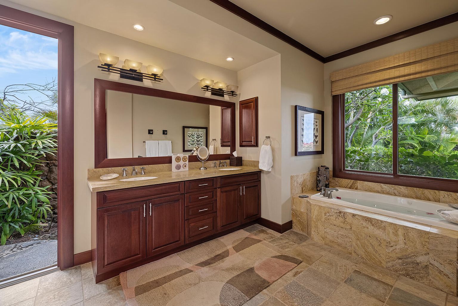 This is a well-appointed primary bathroom featuring a double vanity with dark wood cabinetry and light countertops. A large mirror spans the vanity area, reflecting the neutral-toned walls and recessed lighting. A soaking tub with a marble surround sits beneath a window, offering a view of lush greenery, creating a spa-like atmosphere.