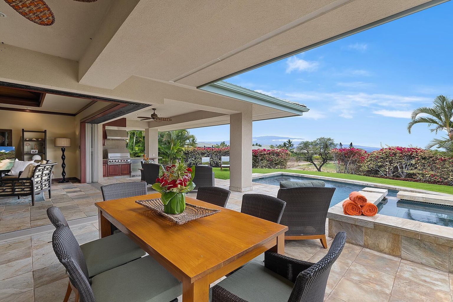 This image showcases a luxurious outdoor patio area, perfect for entertaining or relaxing. The space features a wooden dining table with seating, a built-in grill, and a stunning view of a pool and lush landscaping. The patio seamlessly blends indoor and outdoor living, creating an inviting and upscale atmosphere.