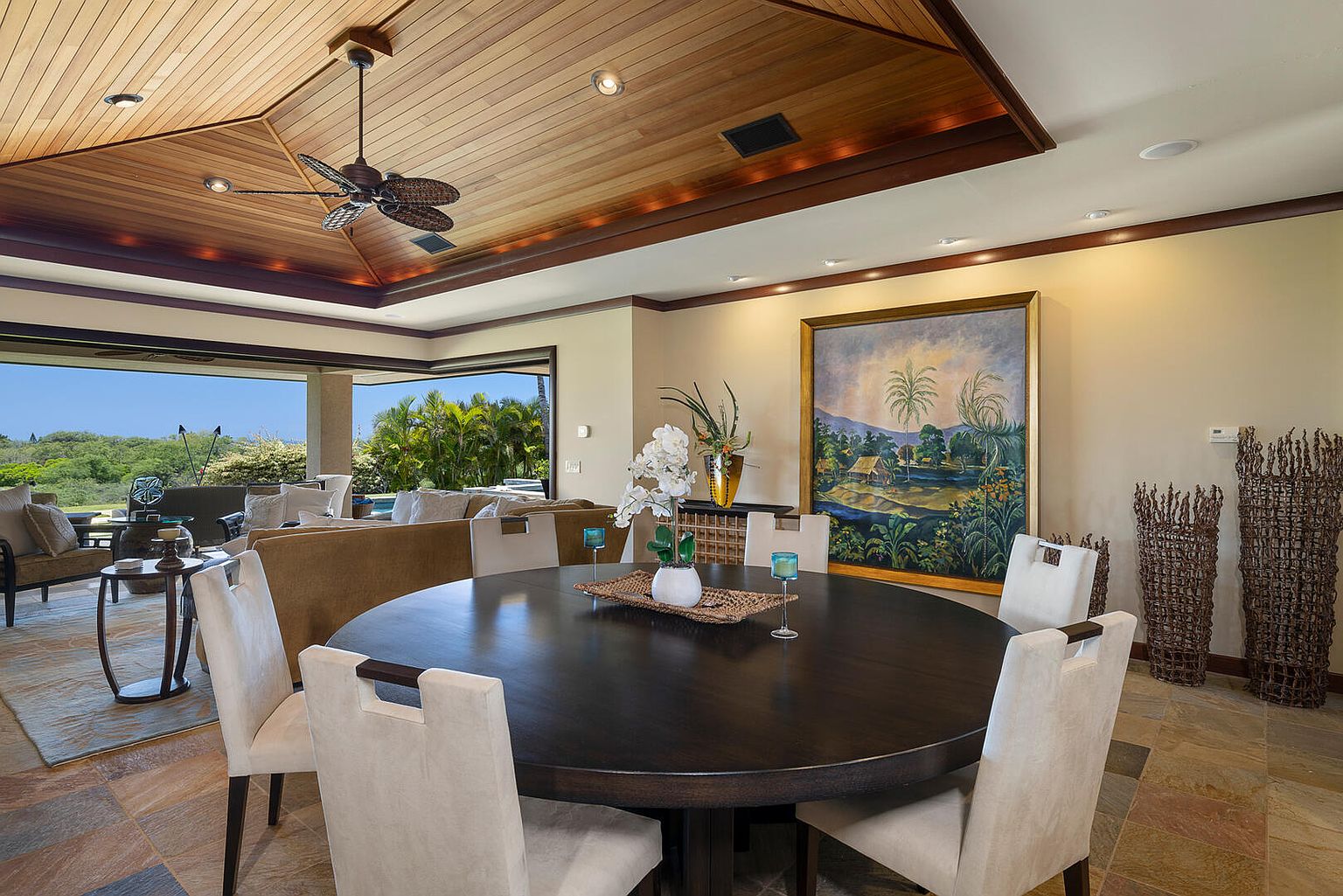 This interior shot showcases a dining room with a large, round dark wood table surrounded by light-colored upholstered chairs. A painting hangs on the wall, and the room opens to an outdoor living area with lush greenery. The space is well-lit and exudes a sense of luxury and comfort.