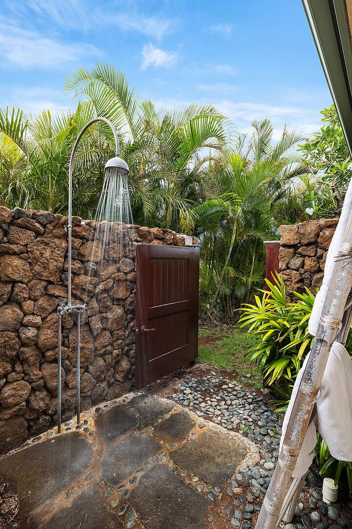 This image showcases an outdoor shower area, enclosed by natural stone walls and lush tropical foliage. A wooden door provides privacy, while a stone-paved shower floor and pebble ground cover add to the natural aesthetic. The showerhead is actively running, creating a refreshing and inviting atmosphere.