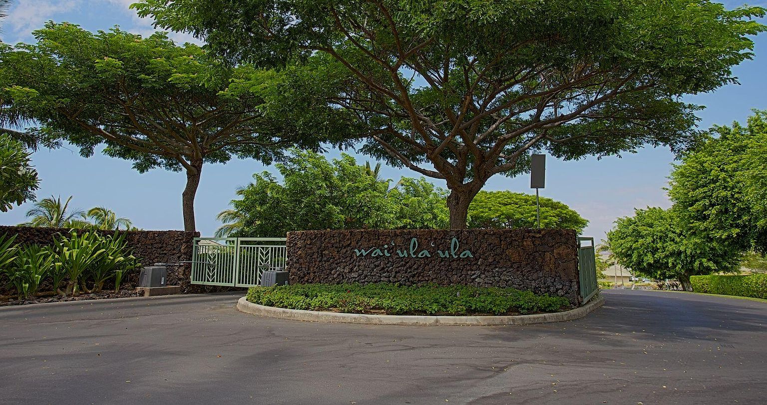 The image showcases the entryway to a residential community, featuring a stone wall with the community's name, 'Wai'ula'ula,' inscribed on it. Lush greenery surrounds the wall, and mature trees provide shade, creating a welcoming and upscale impression. An ornate gate is visible, suggesting a secure and private environment.