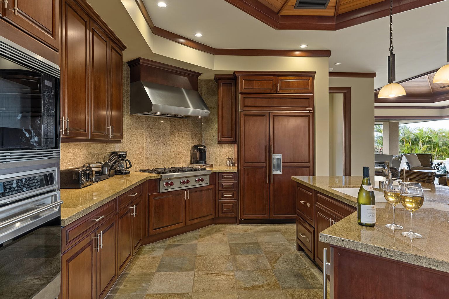 This is a well-appointed kitchen featuring dark wood cabinetry, granite countertops, and stainless steel appliances. The kitchen includes a large island with a sink and wine glasses, suggesting an area for entertaining. The flooring is a neutral-toned tile, and the overall impression is one of luxury and functionality.