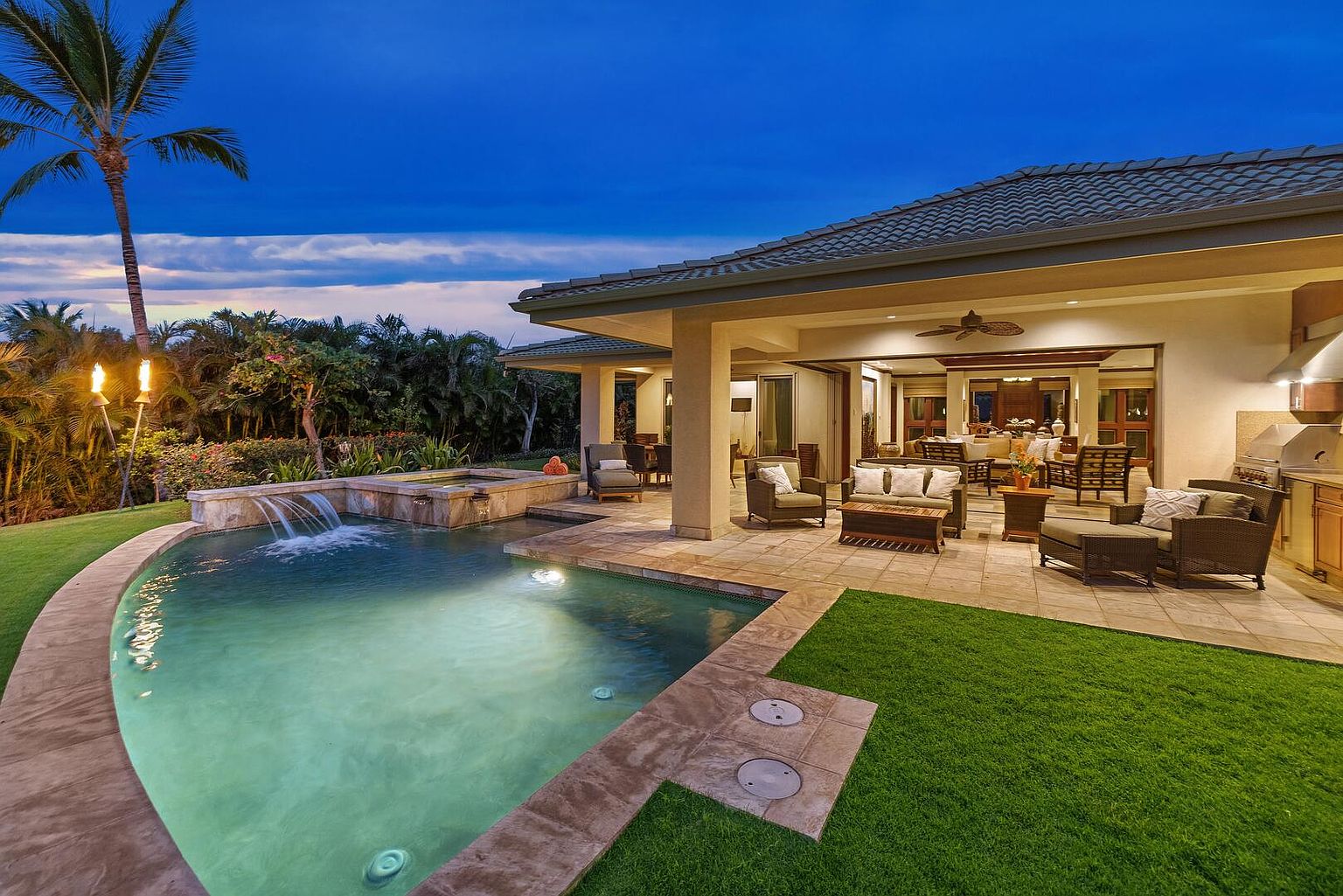 This image showcases a luxurious backyard featuring a pool and spa area. The pool has a waterfall feature and is surrounded by a stone patio that transitions into a well-manicured lawn. Adjacent to the pool is an outdoor living space with comfortable seating, suggesting an ideal setting for relaxation and entertainment. The perspective is from the side, capturing the pool, patio, and part of the house.