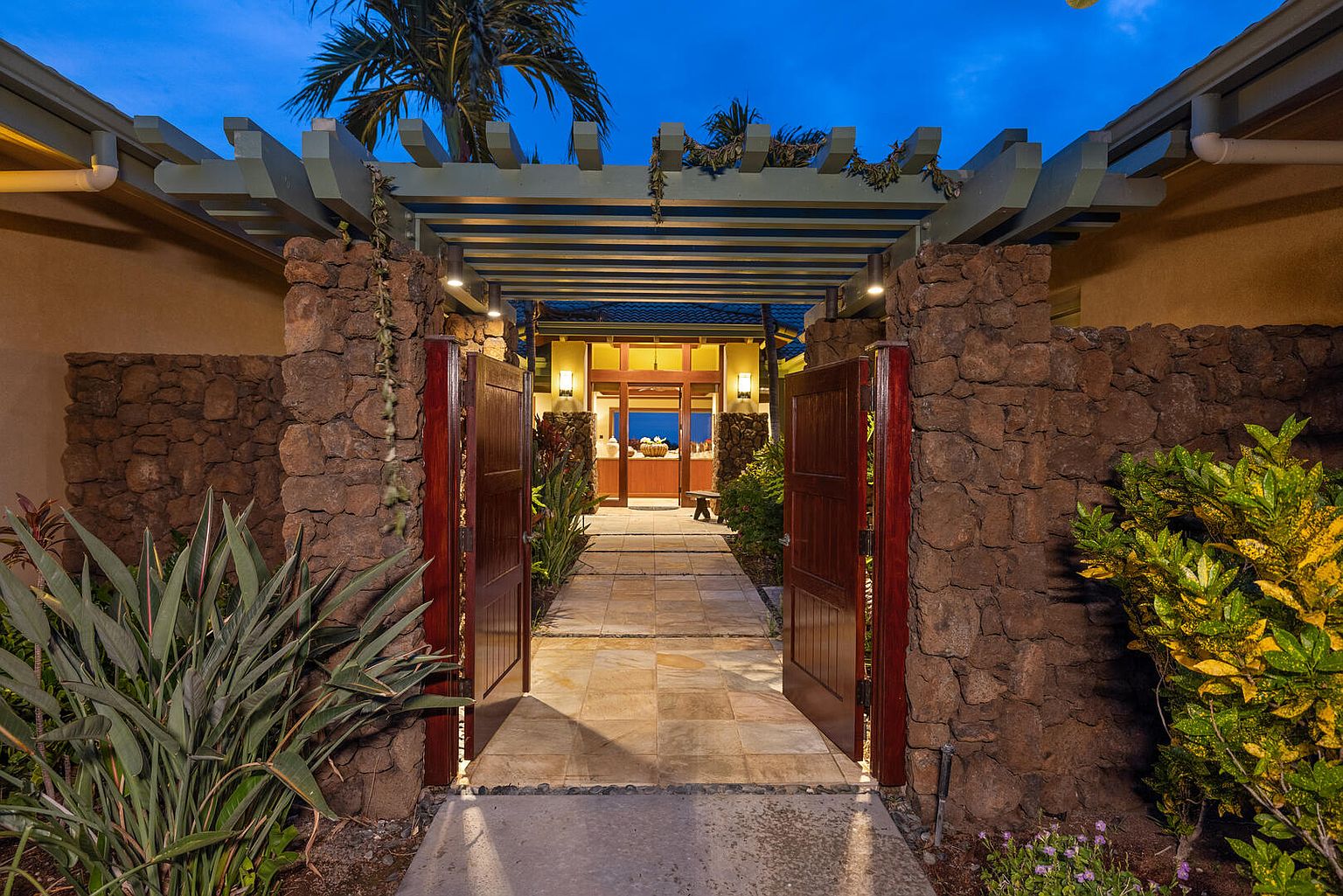 This image showcases a grand entryway featuring an open wooden gate flanked by stone pillars and lush greenery. A pergola overhead adds architectural interest, while the pathway leads to a warmly lit interior, hinting at a welcoming home. The overall impression is one of luxury and tranquility.