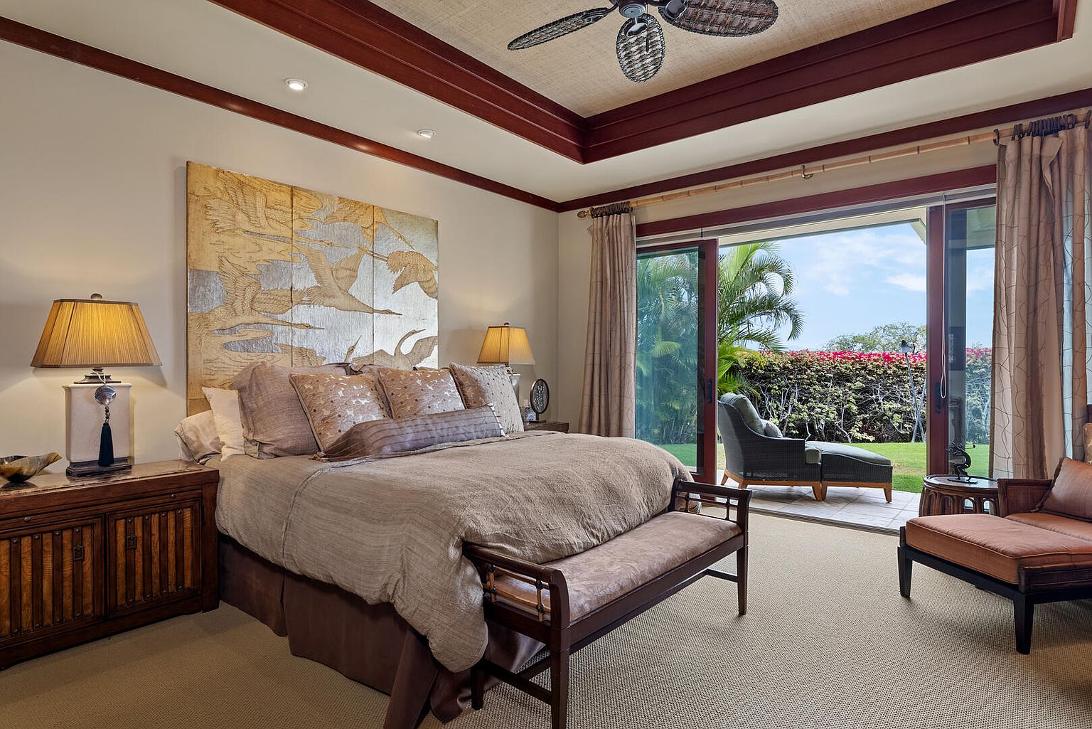 This is a primary bedroom featuring a large bed with a decorative headboard and a bench at the foot. Sliding glass doors lead to an outdoor patio area with lush greenery. The room has a warm and inviting atmosphere, with a ceiling fan and tasteful decor.