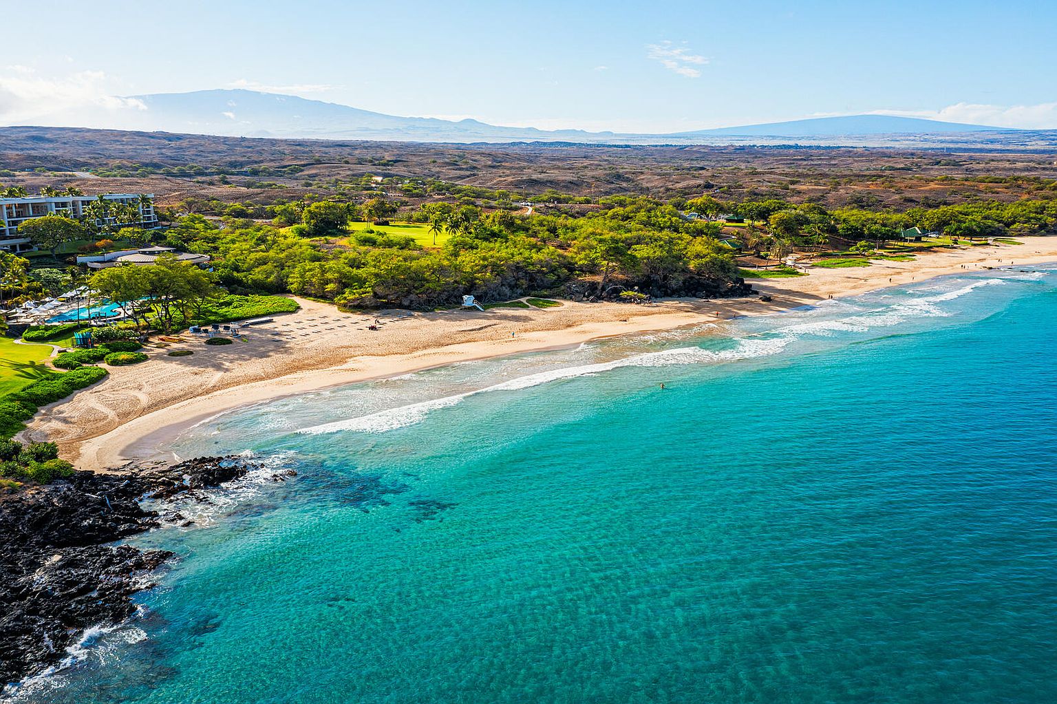 This aerial view showcases a stunning coastal property, featuring a pristine sandy beach meeting crystal-clear turquoise waters. Lush greenery and mature trees border the beach, leading to a luxurious resort with a pool. In the background, rolling hills and mountains provide a scenic backdrop, enhancing the property's appeal and sense of exclusivity.