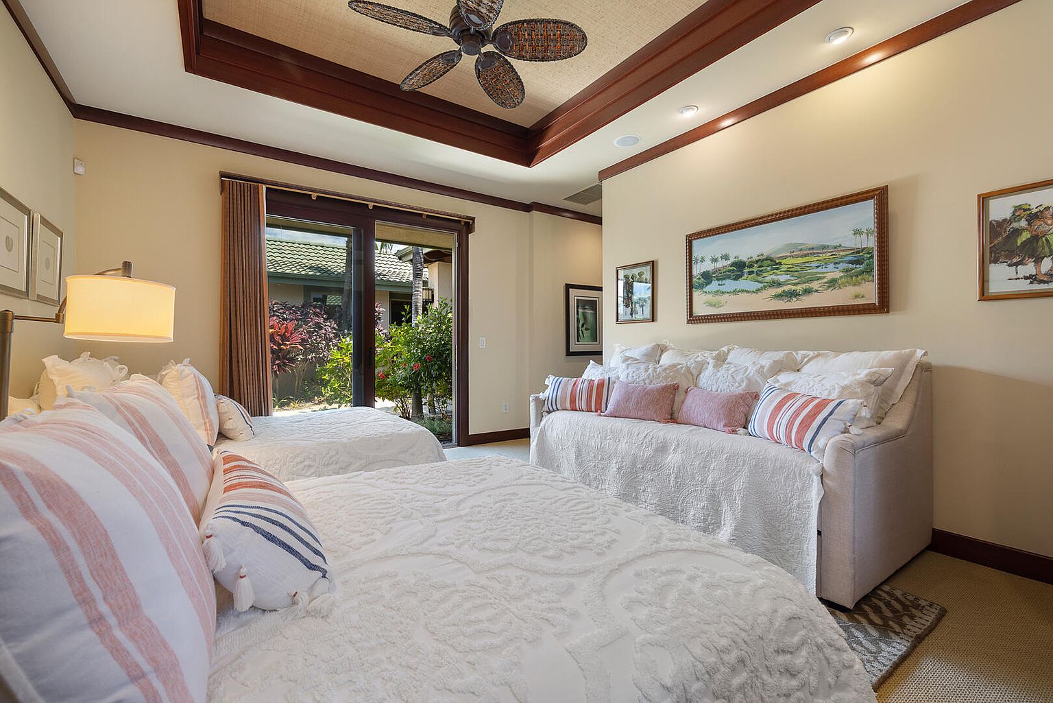 This is a cozy guest bedroom featuring two twin beds and a daybed, all adorned with white patterned bedding and decorative pillows. The room has a warm, inviting ambiance with neutral-toned walls, dark wood trim, and artwork displayed on the walls. A sliding glass door provides access to the outside, bringing in natural light and a view of the garden.