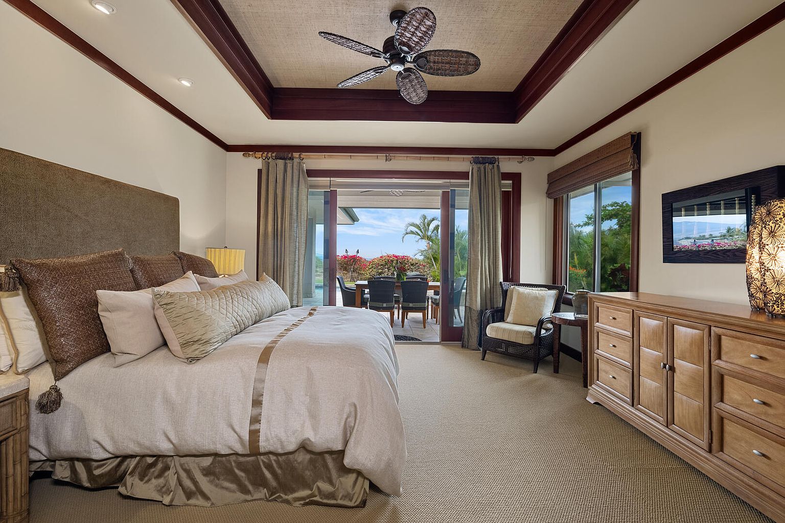 This is a well-appointed primary bedroom featuring a large bed with neutral-toned linens and decorative pillows. The room has a ceiling fan, a dresser, and sliding glass doors that lead to an outdoor patio. The overall impression is luxurious and comfortable, with a focus on relaxation and indoor-outdoor living.