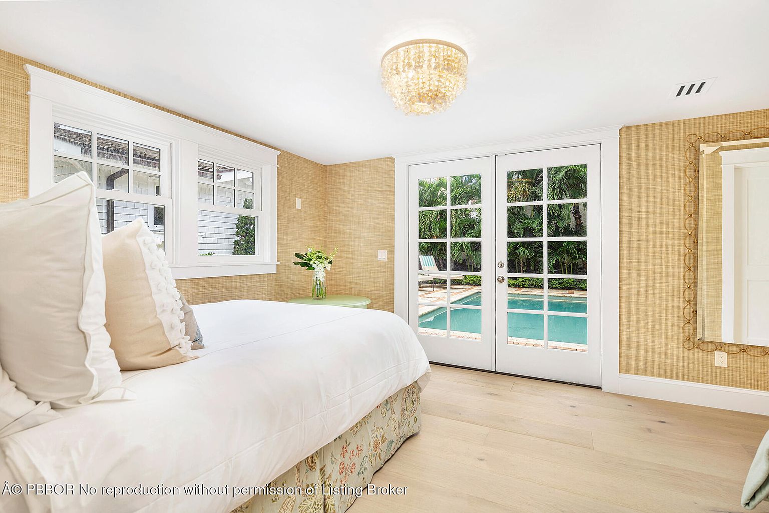 This is a bright and airy bedroom featuring a bed with white linens and decorative pillows, complemented by light wood flooring and textured wallpaper. French doors open to a view of a pool and lush greenery, enhancing the room's tranquil atmosphere. A decorative chandelier adds a touch of elegance.