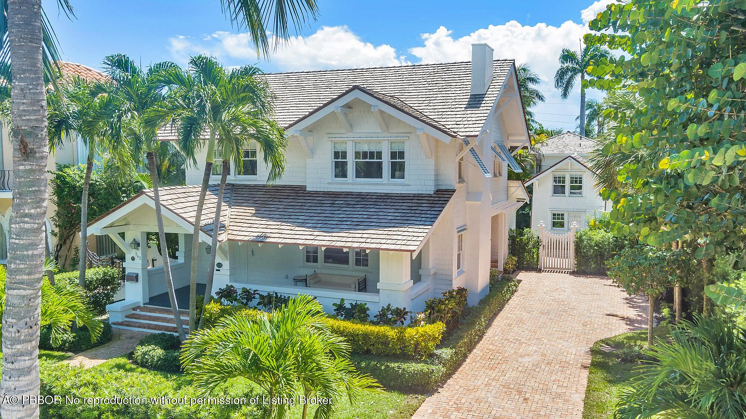 This is a front exterior view of a charming two-story house with a well-manicured lawn and lush landscaping. The house features a light-colored facade, a covered front porch with seating, and a brick driveway leading to a detached garage or guest house in the back. The architectural style evokes a classic coastal or traditional design, enhanced by the surrounding palm trees and greenery.