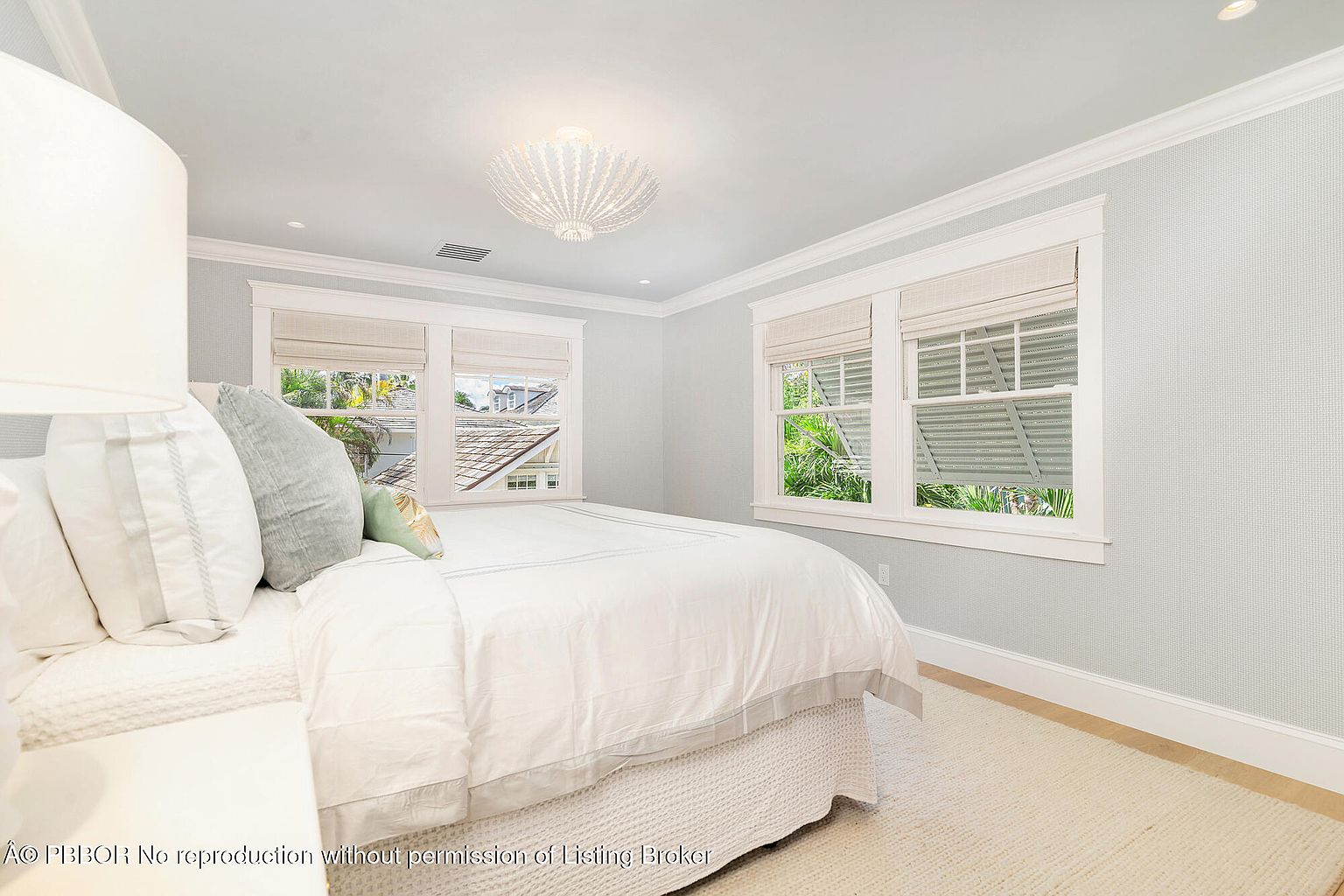 This is a bright and airy bedroom featuring a bed with white linens and decorative pillows. Two windows with woven shades allow natural light to fill the room, complemented by a stylish ceiling light fixture. The walls are painted a soft blue, creating a calming and inviting atmosphere.