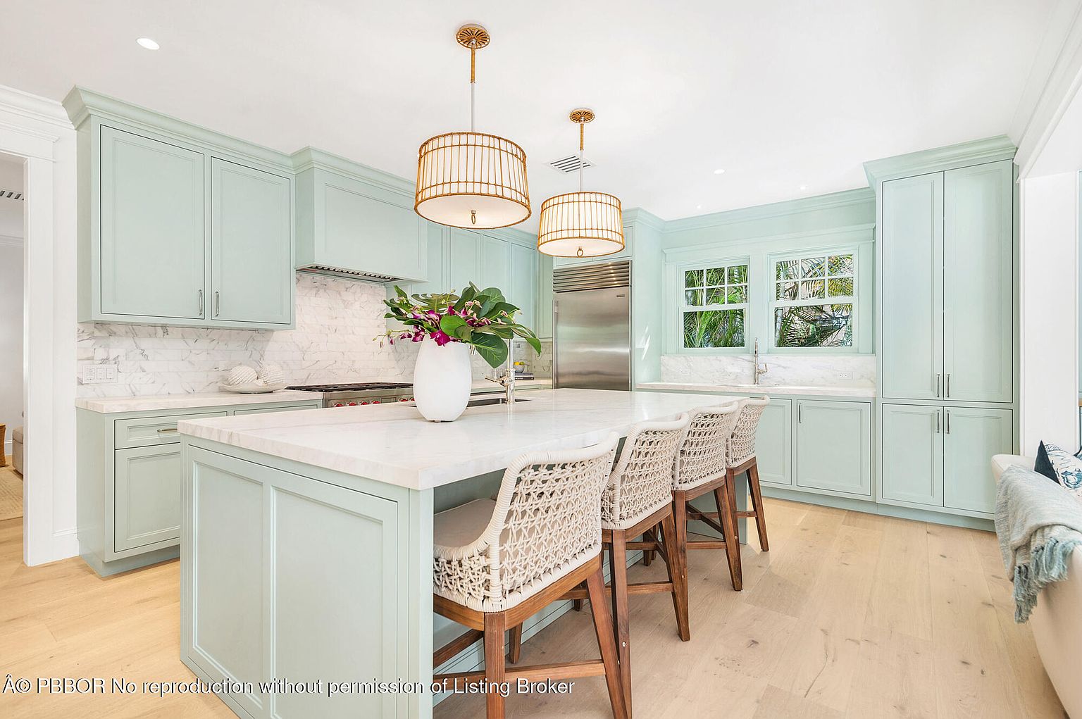 This is a bright and airy kitchen featuring light green cabinetry, marble countertops and backsplash, and stainless steel appliances. A large island with seating is the focal point, illuminated by pendant lighting. The space is clean, modern, and inviting, perfect for both cooking and entertaining.
