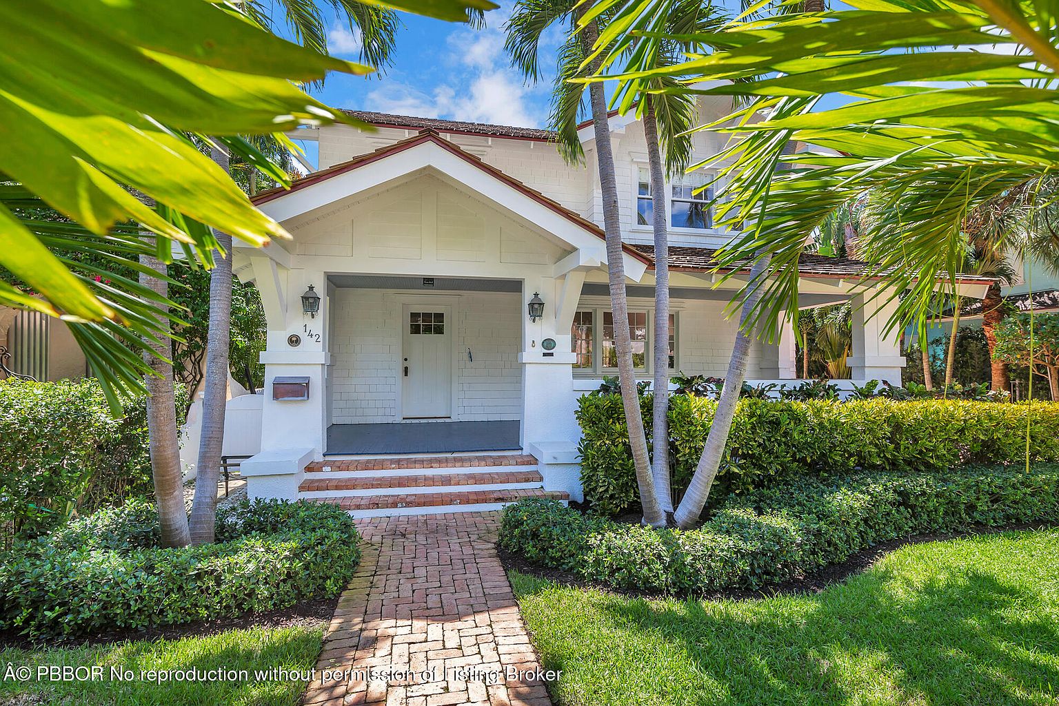 The image showcases the front exterior of a charming two-story house, framed by lush palm trees. A brick pathway leads to the covered entryway, which features a white door and classic architectural details. The well-maintained lawn and landscaping enhance the property's curb appeal, creating a welcoming and inviting atmosphere.