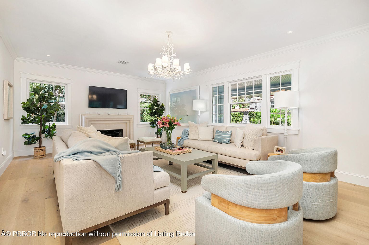 This is a bright and airy living room featuring a neutral color palette. The room is furnished with two sofas, two armchairs, a coffee table, and a decorative chandelier. Large windows provide ample natural light and views of the outdoors, creating a welcoming and comfortable atmosphere.