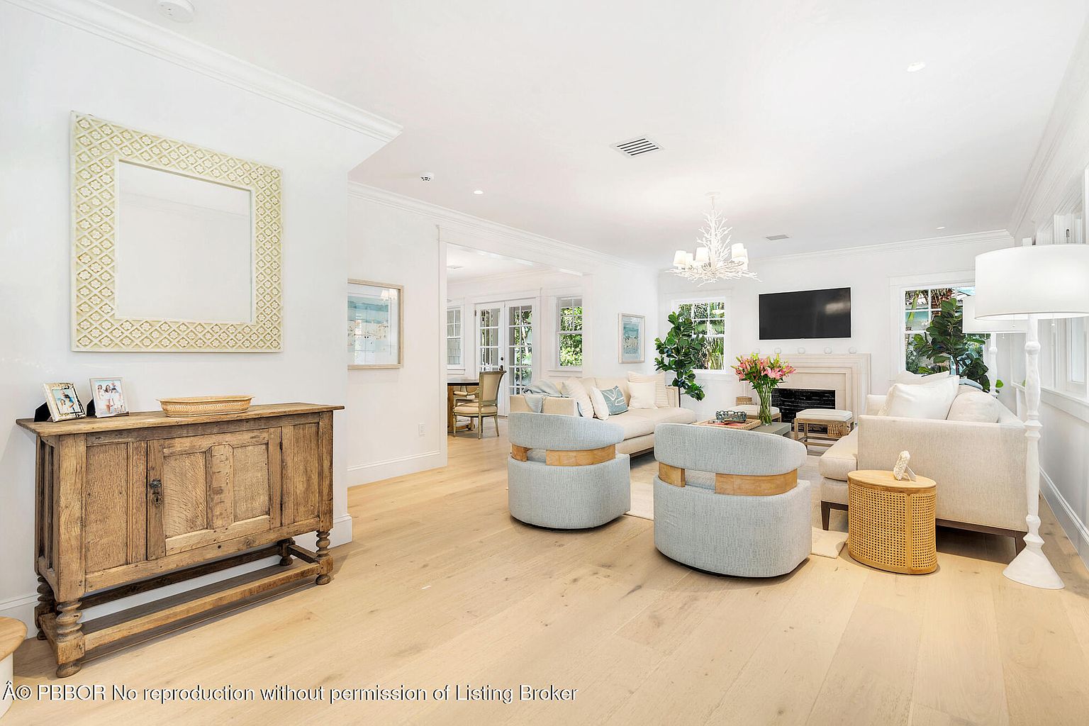 This is a bright and airy living room featuring light hardwood floors, white walls, and ample natural light. The room is furnished with two light blue armchairs, a beige sofa, and a wooden cabinet with a decorative mirror above it. The overall style is elegant and inviting, creating a comfortable and sophisticated living space.
