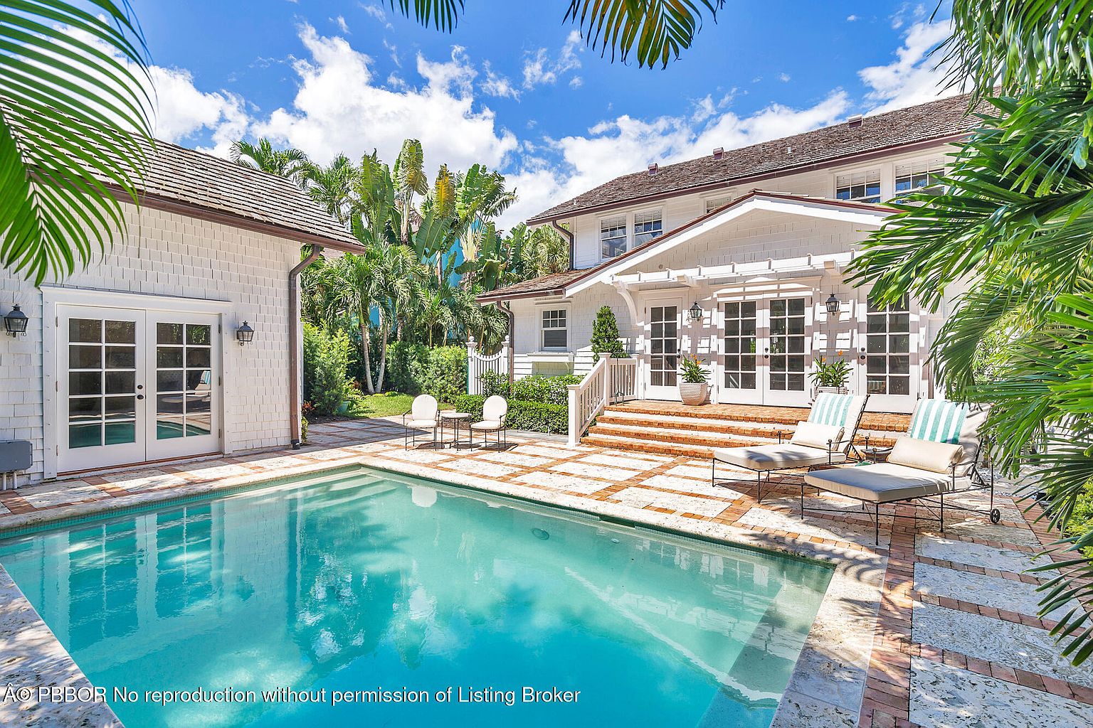 This image showcases a luxurious backyard pool area, featuring a pristine turquoise pool surrounded by a brick-patterned patio. The main house and a smaller outbuilding are visible, both with white exteriors and dark roofs, complemented by lush tropical landscaping. Two lounge chairs are positioned near the pool, inviting relaxation and leisure.