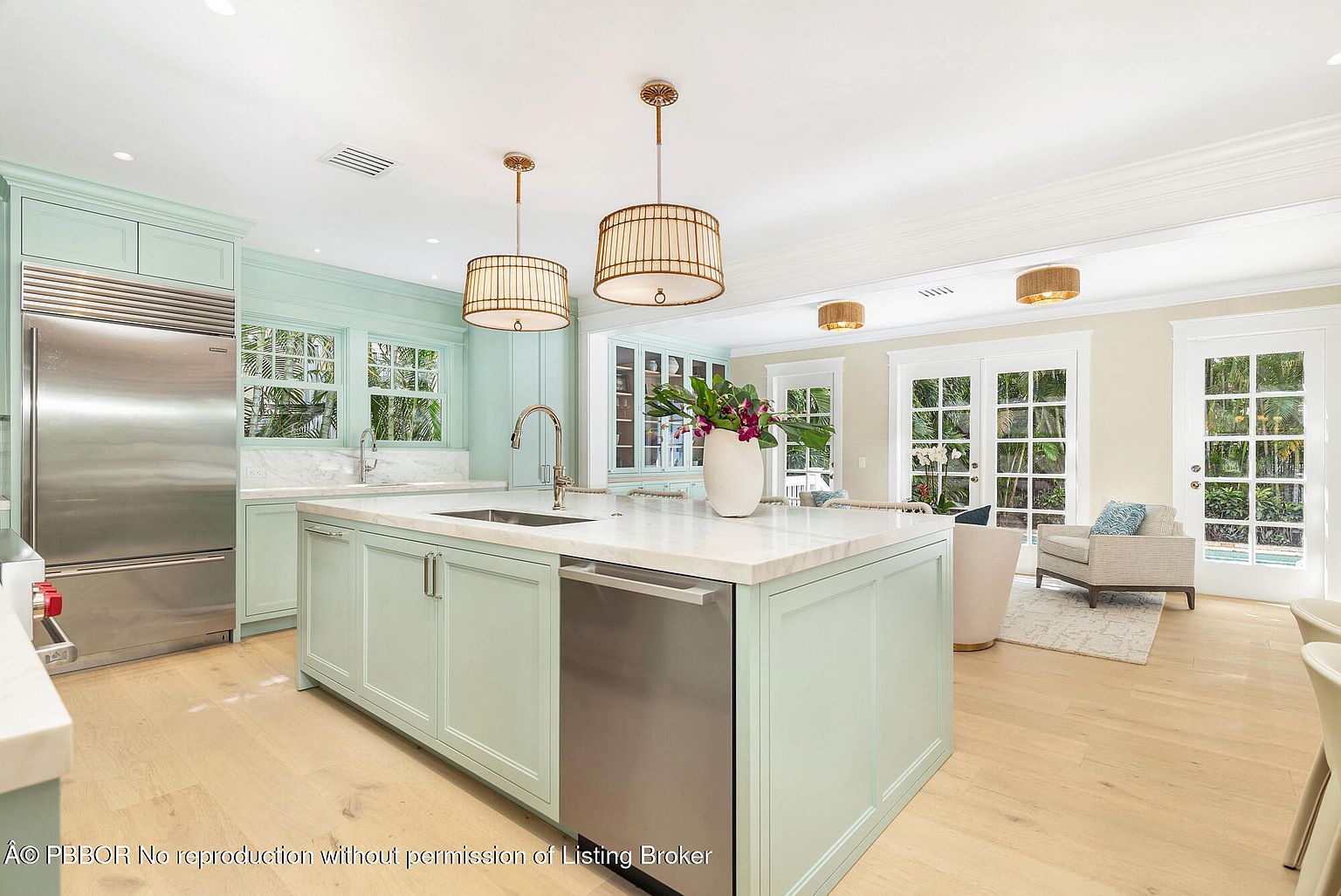 This is a bright and airy kitchen featuring light green cabinetry, stainless steel appliances, and a large island with a white countertop. The kitchen has a modern and elegant design, with pendant lighting and large windows that bring in natural light. The open layout connects the kitchen to a living area, creating a welcoming and spacious atmosphere.