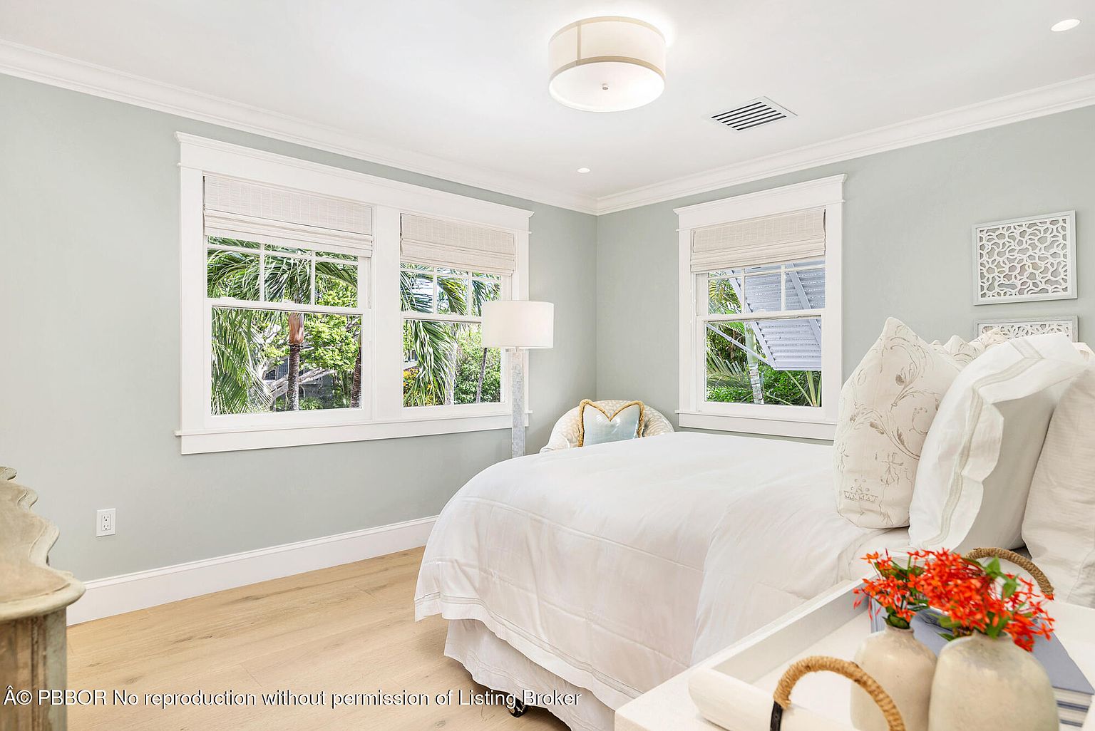 This is a bright and airy primary bedroom featuring a large bed with white linens, complemented by natural light streaming through multiple windows. The room is painted in a soft, neutral tone, and the hardwood floors add warmth. Decorative wall art and a stylish floor lamp enhance the room's aesthetic, creating a serene and inviting atmosphere.