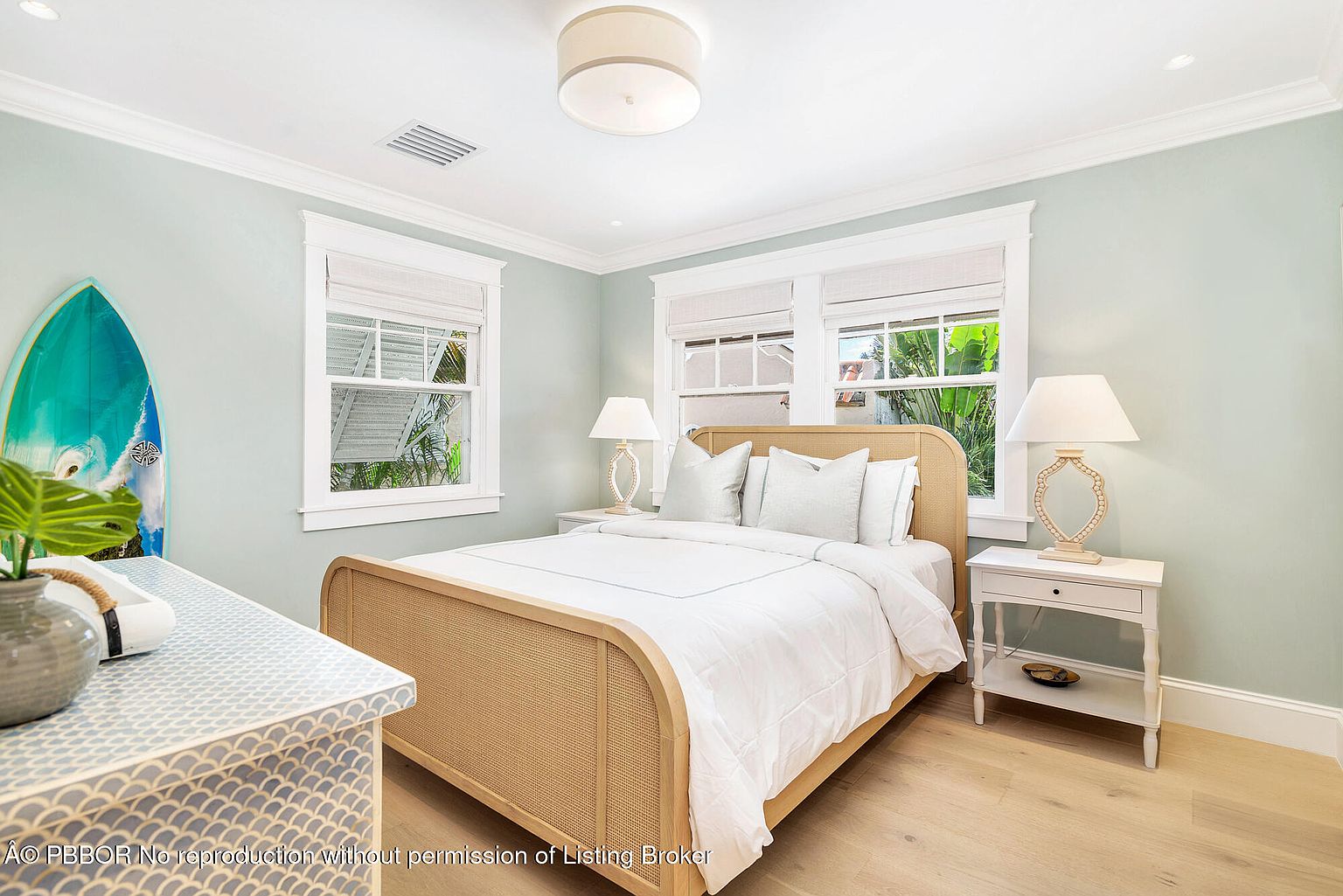 This is a bright and airy guest bedroom featuring a rattan bed with white linens, complemented by matching white nightstands and lamps. Natural light floods the room through the windows, enhancing the light blue walls and light wood flooring. A surfboard adds a touch of coastal charm to the space.