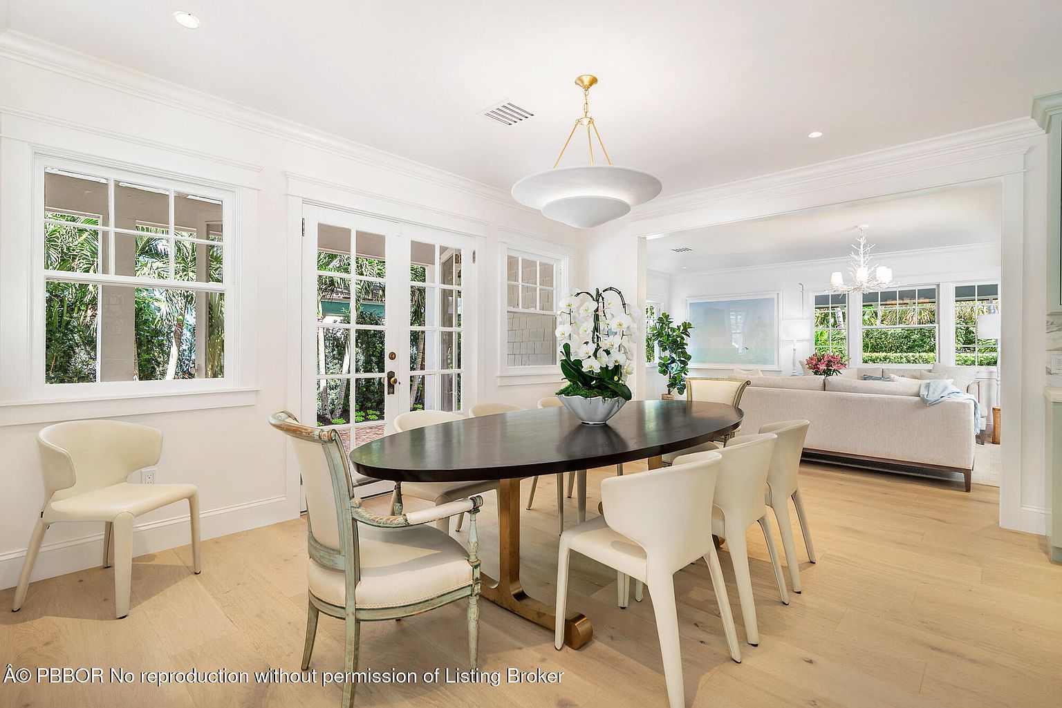 This is an interior shot of a dining room featuring a dark wood oval table surrounded by modern white chairs and two upholstered chairs with light green frames. The room is bright with natural light coming through large windows and French doors, and a modern chandelier hangs above the table. The space opens into a living area, creating an open and airy feel.