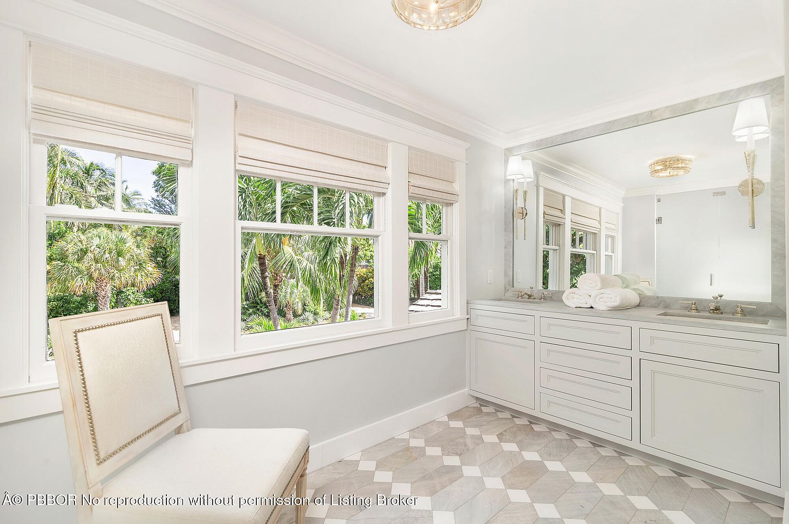 This is a bright and airy bathroom featuring a large window with natural light and a view of lush greenery. The bathroom includes a vanity with white cabinetry, a marble countertop, and a large mirror. The flooring is a patterned tile, and a decorative chair adds a touch of elegance to the space.