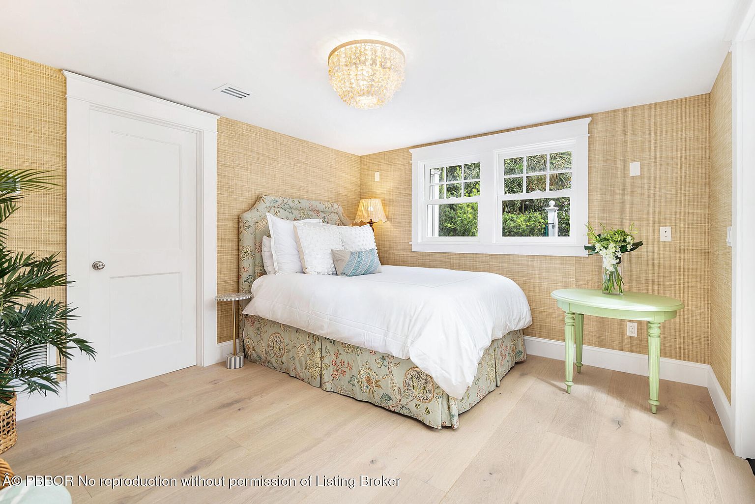 This is a cozy bedroom featuring a floral-patterned bed skirt and headboard, complemented by white bedding and decorative pillows. The walls are covered in a textured wallpaper, and natural light streams in through a window. A light green side table with a vase of flowers adds a touch of freshness to the room.