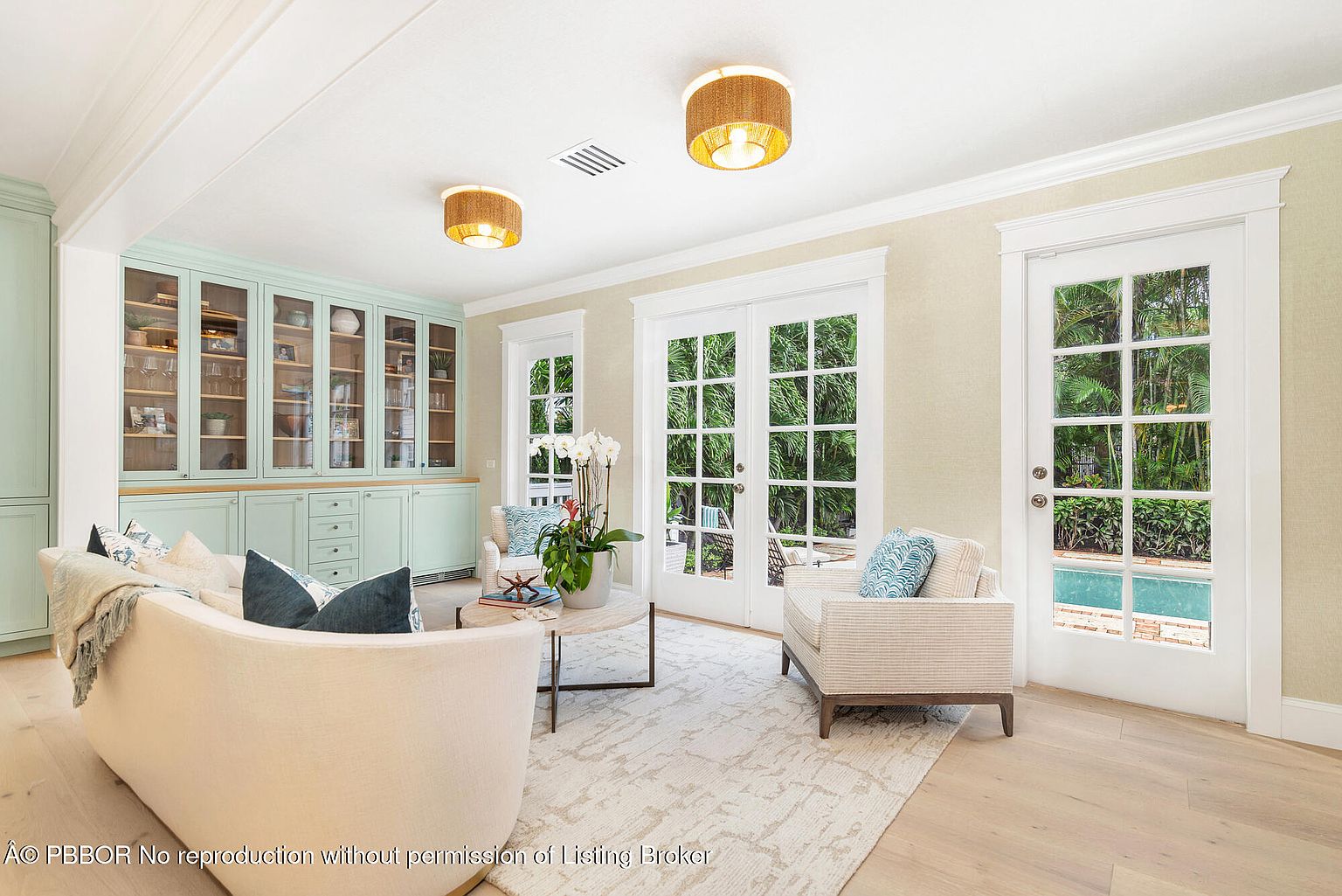 This is a bright and airy living room featuring a curved sofa with throw pillows, a light-colored area rug, and built-in cabinetry in a soft blue hue. French doors provide ample natural light and a view of the outdoor pool area. The room exudes a relaxed and elegant atmosphere.