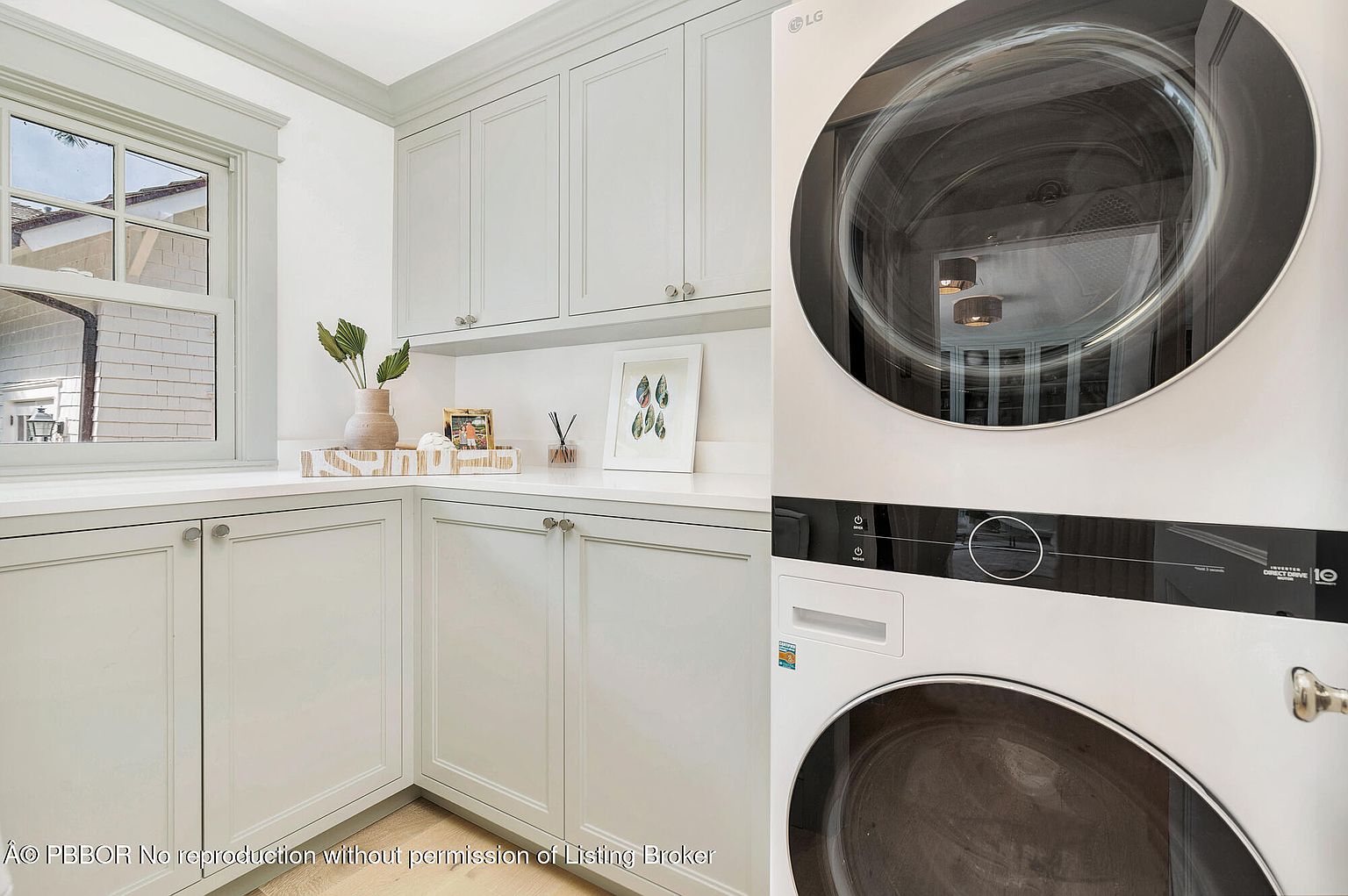This is a well-organized laundry room featuring stacked LG washer and dryer units. The room has light gray cabinetry with silver hardware and a white countertop, providing ample space for folding and sorting laundry. A window provides natural light, and decorative items add a touch of style to the functional space.