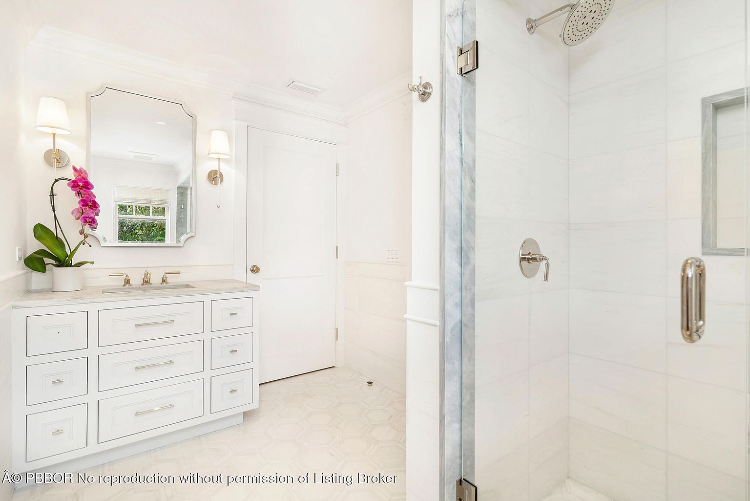 This is a bright and clean primary bathroom featuring a white vanity with marble countertop, a large mirror flanked by sconces, and a glass-enclosed shower with white tile. The floor is tiled in a light-colored geometric pattern, and the overall aesthetic is modern and luxurious. The perspective is from the doorway, showcasing the vanity and shower area.