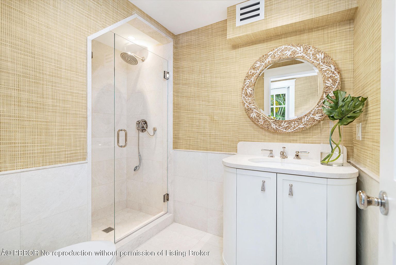 This is a well-lit bathroom featuring a glass-enclosed shower with a rainfall showerhead and a handheld sprayer. The vanity has a white countertop and cabinetry, complemented by a round mirror with a decorative frame. The walls are covered in a textured wallpaper, adding warmth to the space.