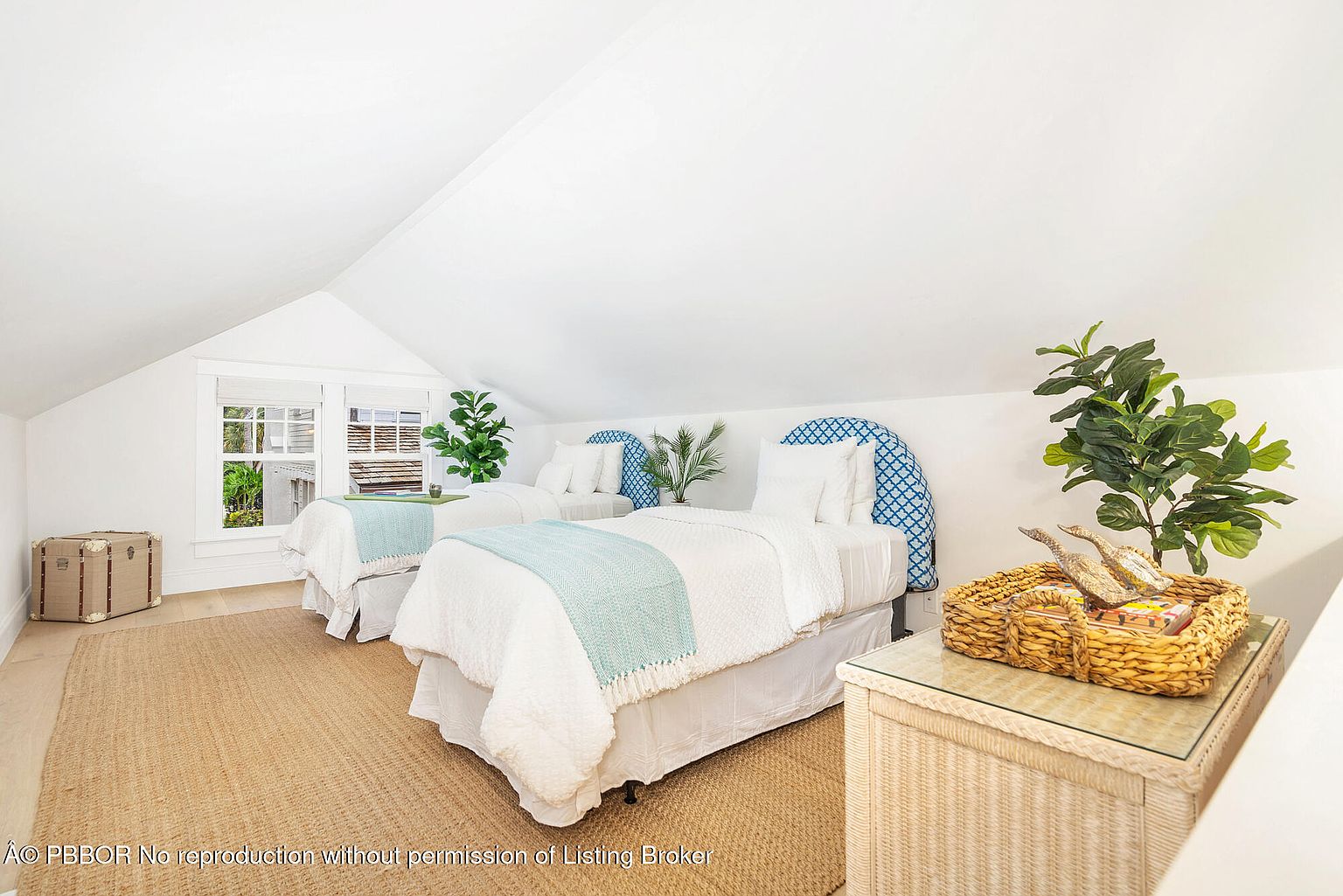 This is a bright and airy guest bedroom featuring two twin beds with white bedding and blue patterned headboards. A woven rug covers the floor, and a wicker nightstand with a glass top sits beside one of the beds. The room has a cozy, inviting feel, enhanced by natural light from the window and the presence of indoor plants.