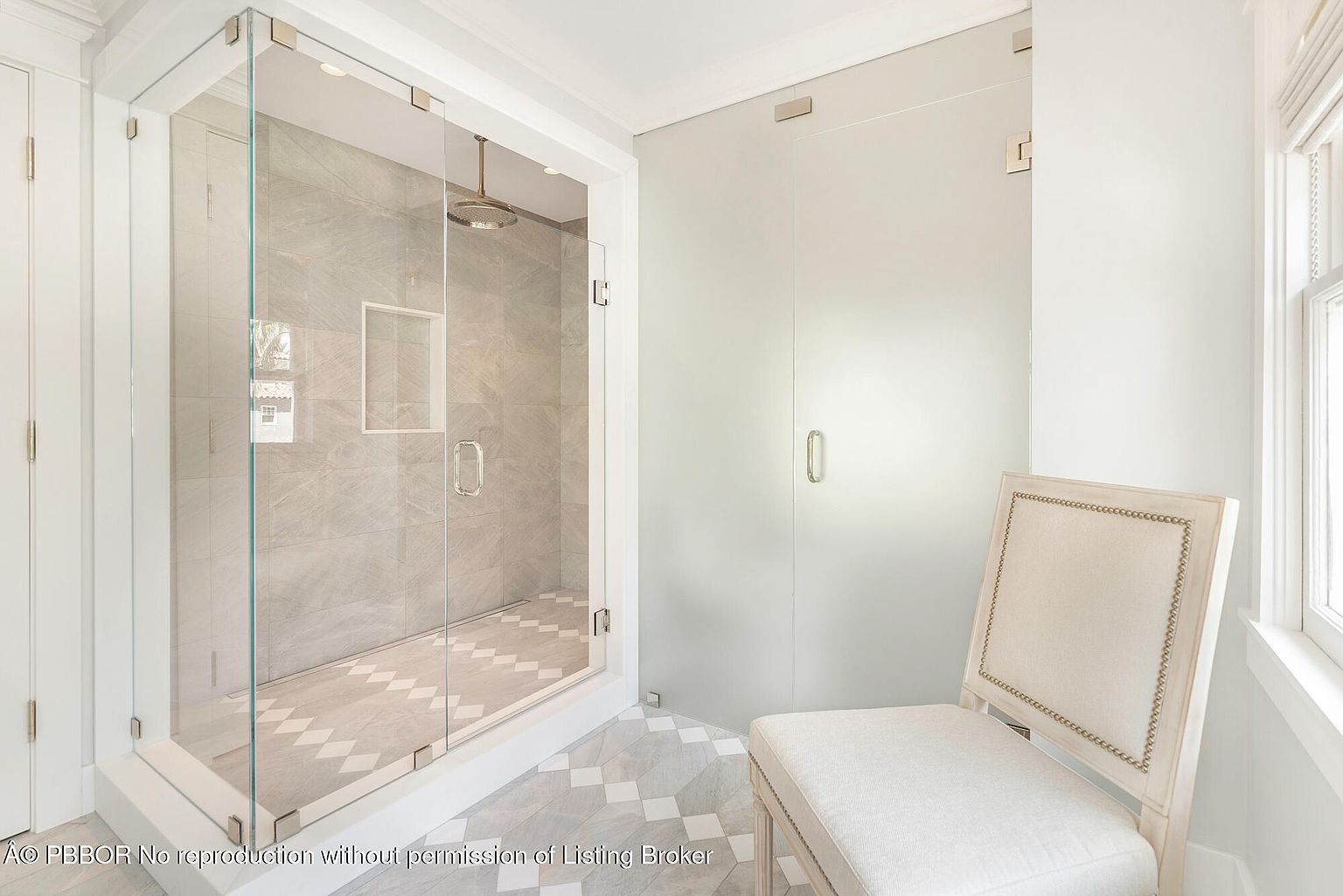This is a bright and modern primary bathroom featuring a glass-enclosed shower with gray tile and a built-in niche. The floor is tiled in a geometric pattern, and a frosted glass door provides privacy for a separate area. A decorative chair sits near the window, adding a touch of elegance to the space.