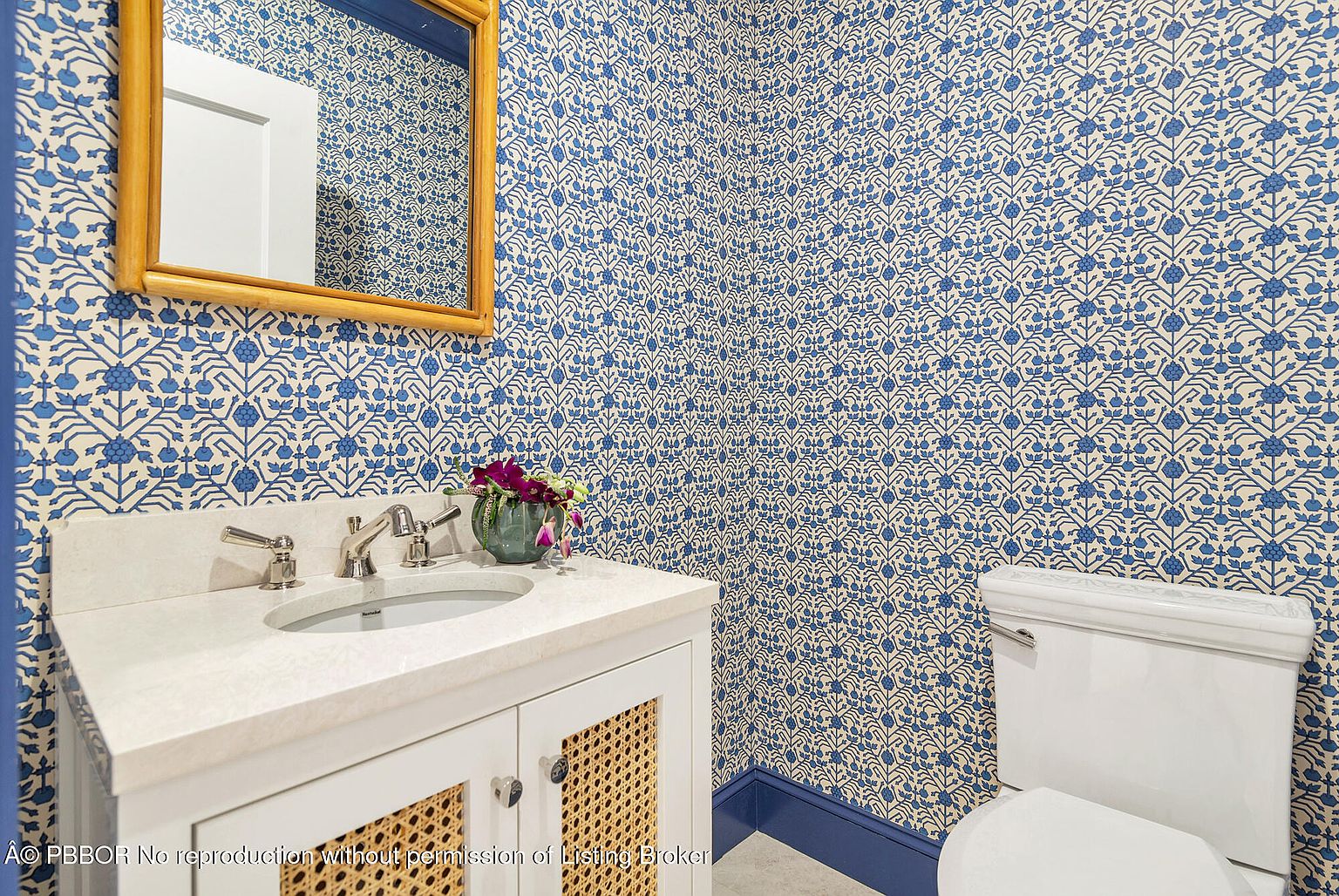 This is a charming guest bathroom featuring a blue and white patterned wallpaper that adds a touch of elegance. The vanity has a white countertop and cabinet with woven door panels, complemented by a gold-framed mirror. A toilet is visible to the right, completing the functional space.