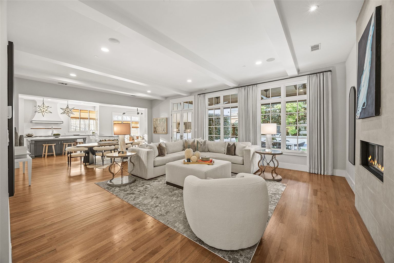 This is a bright and spacious living room featuring hardwood floors, a large sectional sofa, and an ottoman. The room is well-lit with natural light from large windows and recessed lighting. A fireplace and a neutral color palette create a warm and inviting atmosphere.