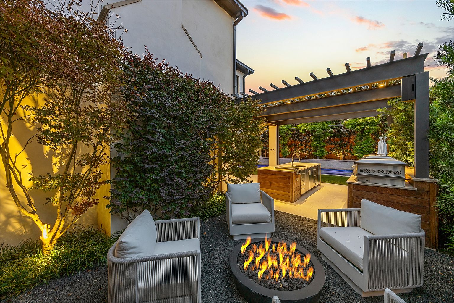 This image showcases a beautifully designed backyard featuring a fire pit surrounded by comfortable seating, creating an inviting outdoor living space. A pergola covers an outdoor kitchen area with a built-in grill and sink, while lush greenery and strategic lighting enhance the ambiance. The overall impression is one of luxury and relaxation, perfect for entertaining or enjoying quiet evenings.