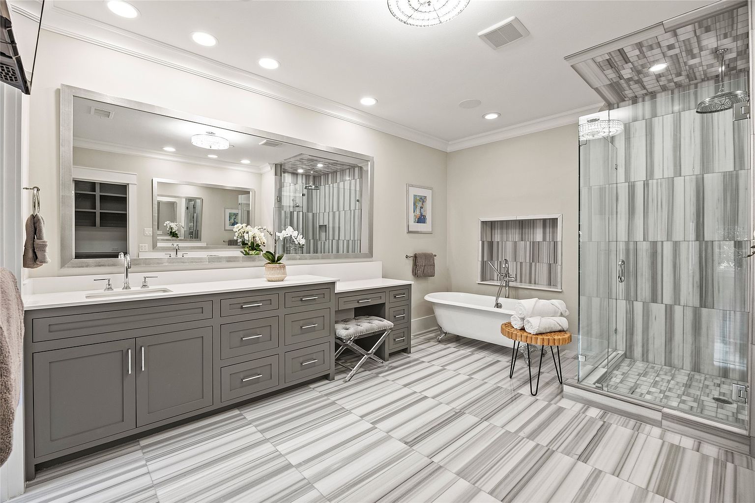 This is a luxurious primary bathroom featuring a large vanity with gray cabinetry and a white countertop, a freestanding bathtub, and a glass-enclosed shower with marble-like tiling. The floor is also tiled with a similar marble pattern, creating a cohesive and elegant design. The room is well-lit with recessed lighting and a decorative chandelier in the shower area, enhancing the spa-like atmosphere.