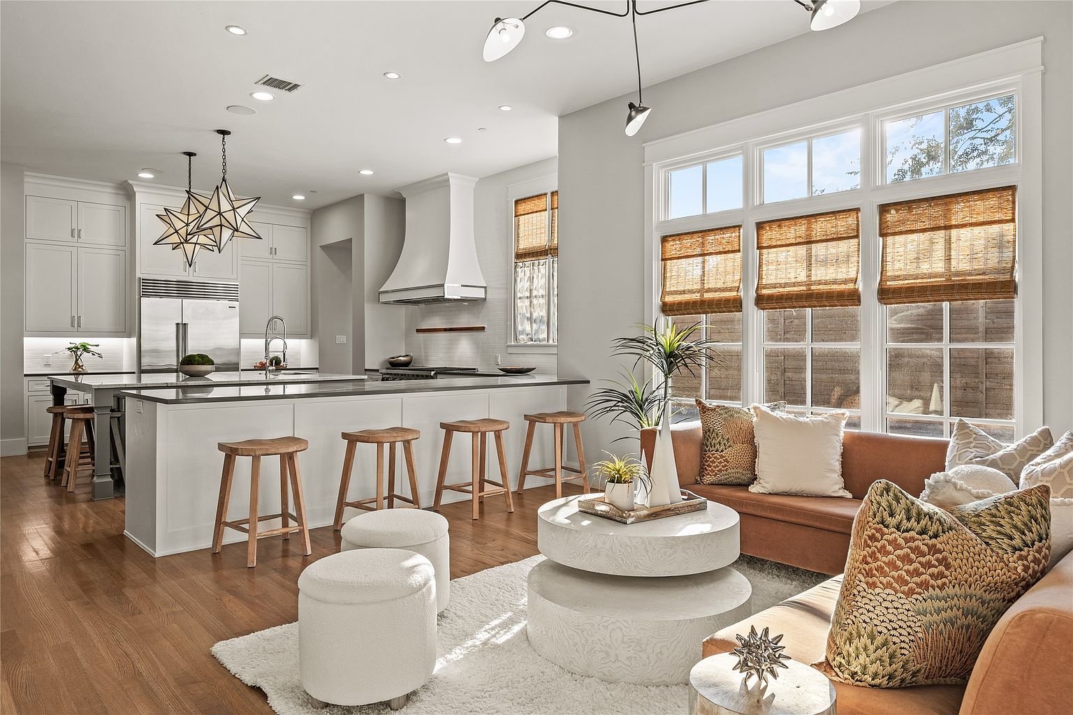 This is a bright and airy living room and kitchen area featuring a modern design with a neutral color palette. The living room area has a comfortable brown leather sofa with decorative pillows, a round white coffee table, and large windows with woven shades. The kitchen features a large island with wooden stools and white cabinets, creating a seamless transition between the two spaces.