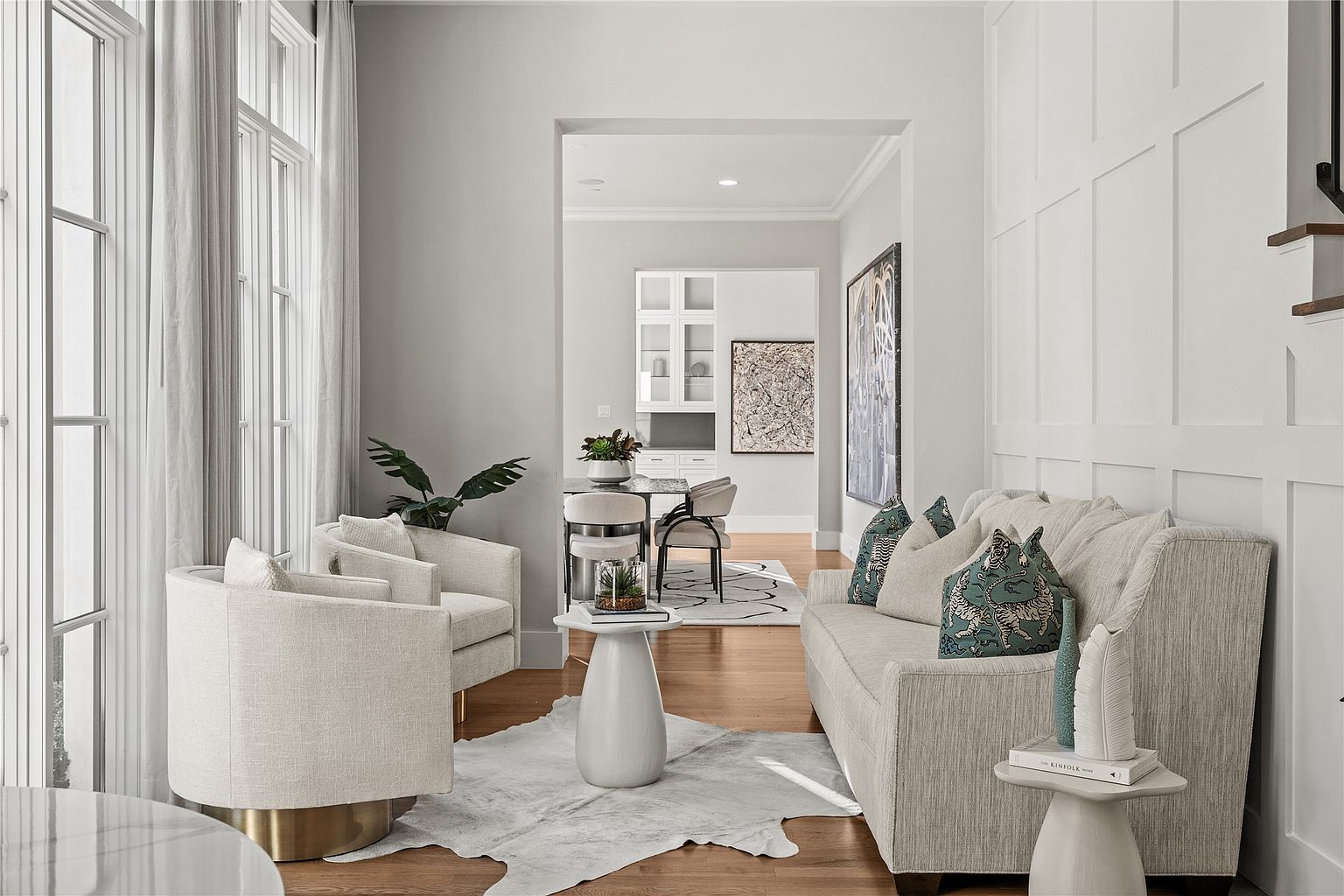 This is an interior shot of a well-lit living room, showcasing a blend of modern and classic design elements. The room features two armchairs, a sofa with decorative pillows, and a white side table, all arranged on a light-colored hide rug over hardwood floors. The color palette is neutral with pops of color from the pillows and artwork, creating a sophisticated and inviting atmosphere.