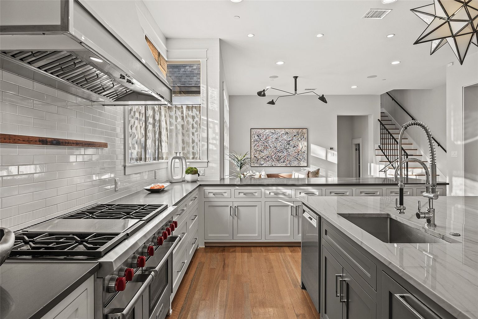 This is a bright and modern kitchen featuring a large island with a marble countertop and stainless steel sink. The kitchen is equipped with high-end appliances, including a professional-grade range with red knobs and a stainless steel vent hood. The hardwood floors and neutral color palette create a warm and inviting atmosphere, while the open layout connects the kitchen to the living area.