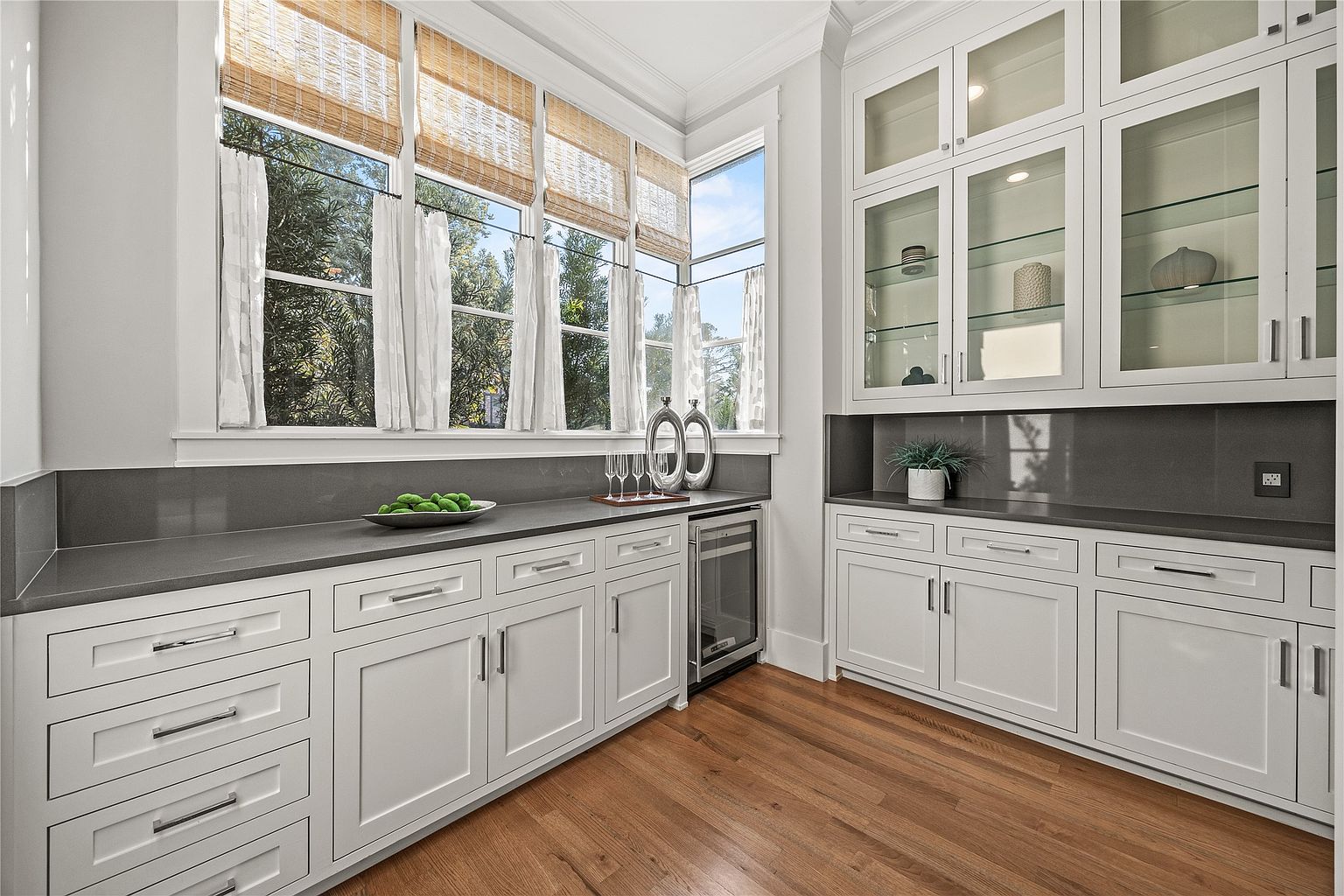 This interior shot showcases a well-organized pantry area with custom white cabinetry, gray countertops, and hardwood flooring. The built-in cabinets feature both drawers and doors, with glass-fronted upper cabinets displaying decorative items. A wine cooler is integrated into the cabinetry, and natural light streams in through a large window, creating a bright and inviting space.