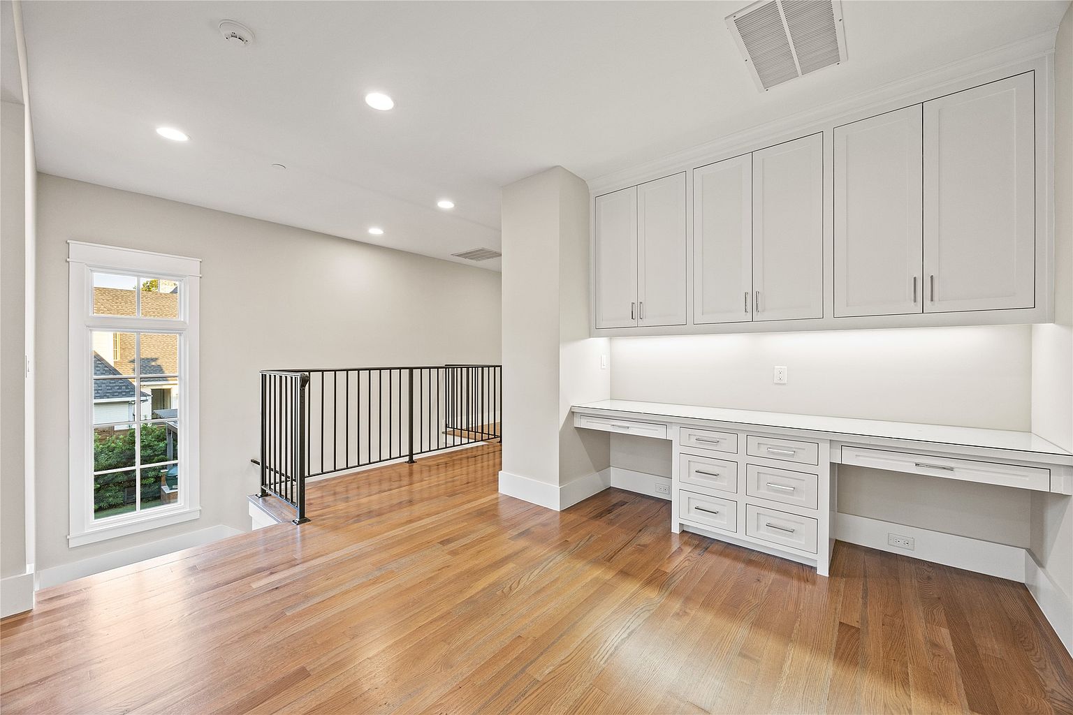 This is an interior shot of a home office or study area. The room features hardwood floors, a built-in desk with white cabinetry and drawers, and upper cabinets for storage. A window provides natural light, and a black railing suggests a landing or hallway area adjacent to the office space.