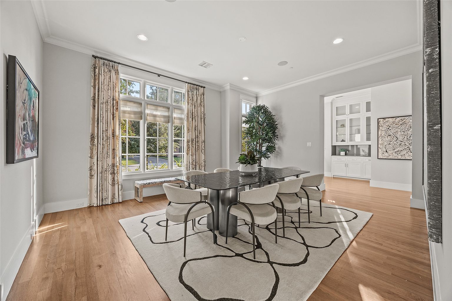 This is an interior shot of a dining room featuring a modern aesthetic. A dark glass-topped dining table is surrounded by six upholstered chairs, all set upon a large area rug with a floral pattern. Natural light floods the room through a large window dressed with patterned curtains, and a built-in cabinet is visible in the background, adding to the room's elegance.