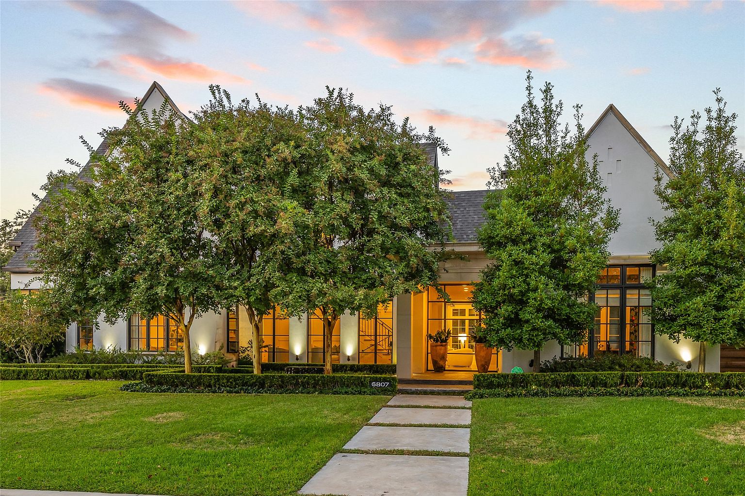 This is a front exterior view of a stately home with a well-manicured lawn and mature trees. The house features a light-colored facade, dark-framed windows, and gabled rooflines, creating a sophisticated and elegant appearance. A stone pathway leads to the entrance, enhancing the curb appeal and inviting feel of the property.
