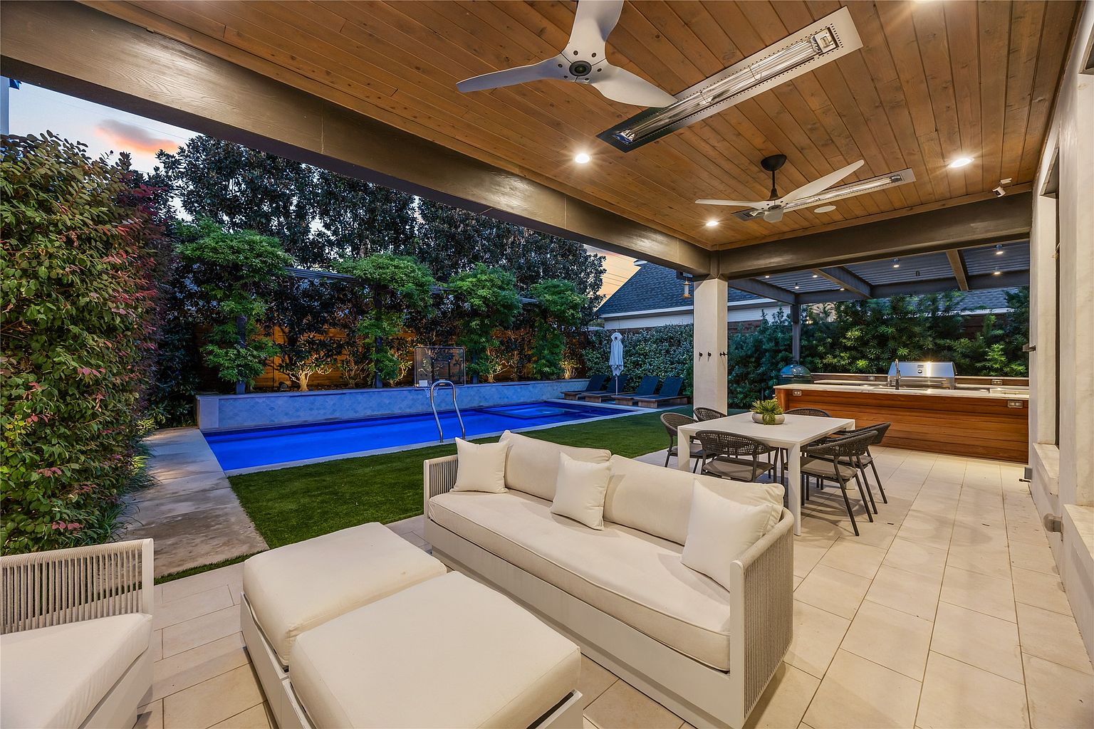 This image showcases a luxurious outdoor patio area, featuring comfortable seating arrangements, including a plush sofa and ottomans. The patio is well-appointed with a dining table and chairs, an outdoor kitchen area, and a view of a beautifully landscaped backyard with a pool. The space is covered by a wooden ceiling with fans and recessed lighting, creating an inviting and sophisticated atmosphere.