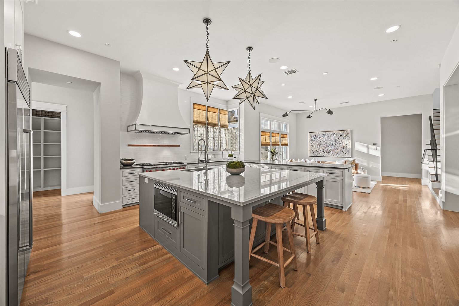 This is a bright and spacious kitchen featuring a large central island with seating, pendant star-shaped lighting, and hardwood floors. The kitchen is equipped with modern appliances and has a clean, contemporary design with white walls and gray cabinetry. The open layout connects to a living area, creating a seamless flow.
