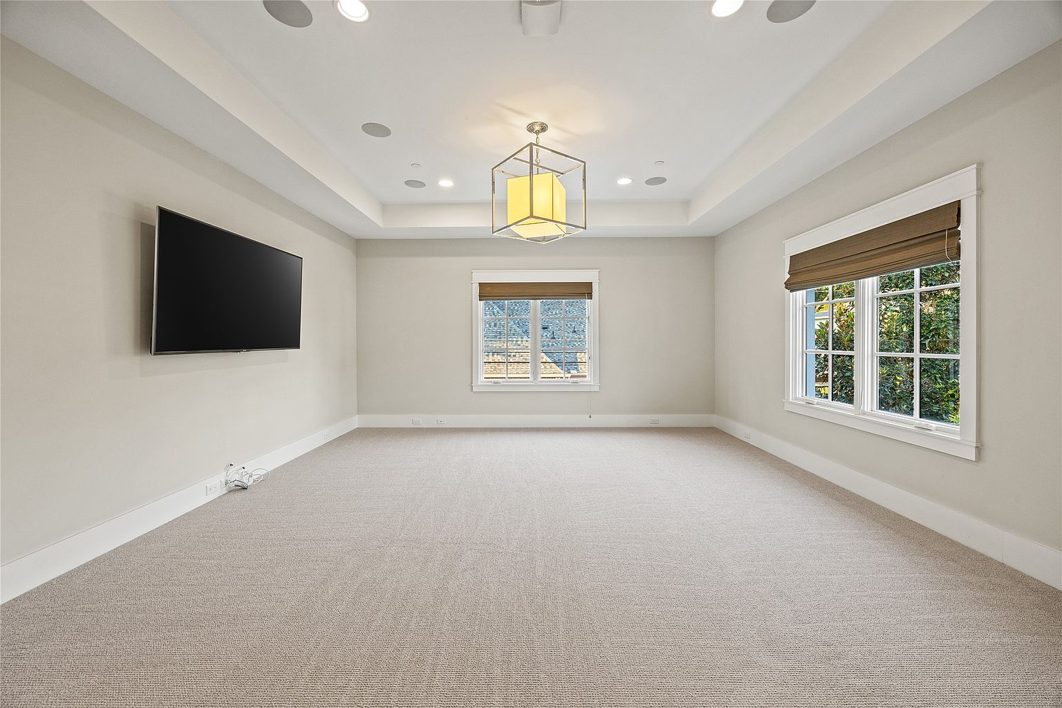 This is a spacious living room featuring neutral-toned walls and carpet, creating a serene atmosphere. The room is well-lit with recessed lighting and a decorative chandelier, complemented by natural light streaming through two windows with woven shades. A large flat-screen TV is mounted on the wall, suggesting a comfortable entertainment space.