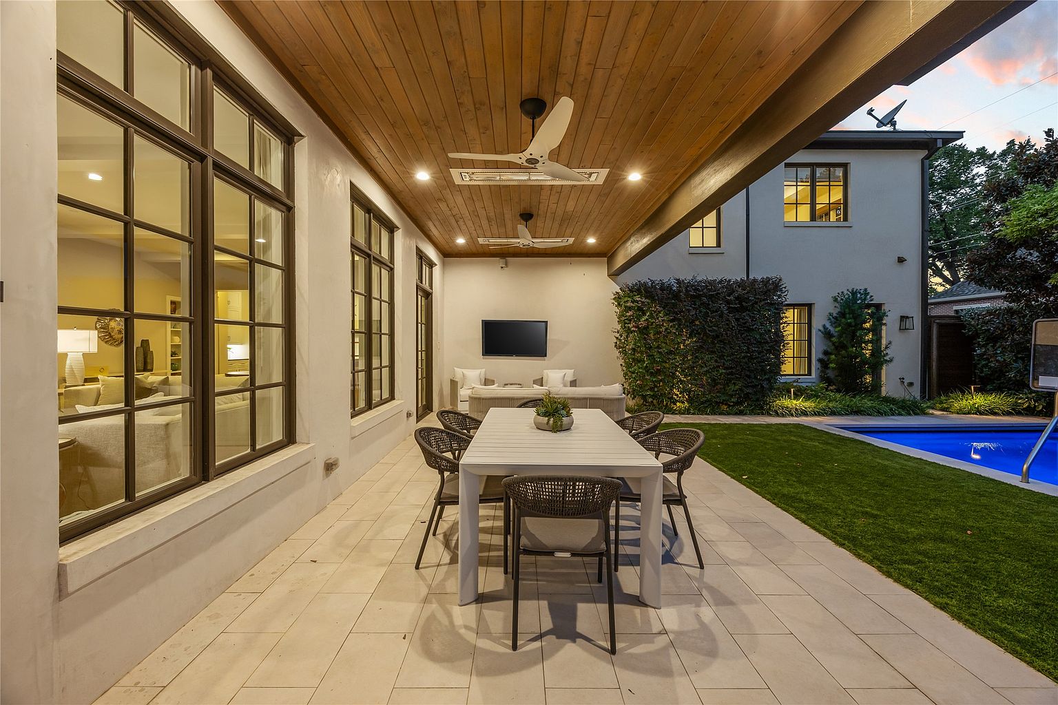 This image showcases a beautifully designed outdoor patio area, featuring a long white dining table with woven chairs under a wooden-paneled ceiling with ceiling fans. The patio is adjacent to a well-manicured lawn and a sparkling blue pool, with a modern home visible in the background, creating an inviting and luxurious outdoor living space.
